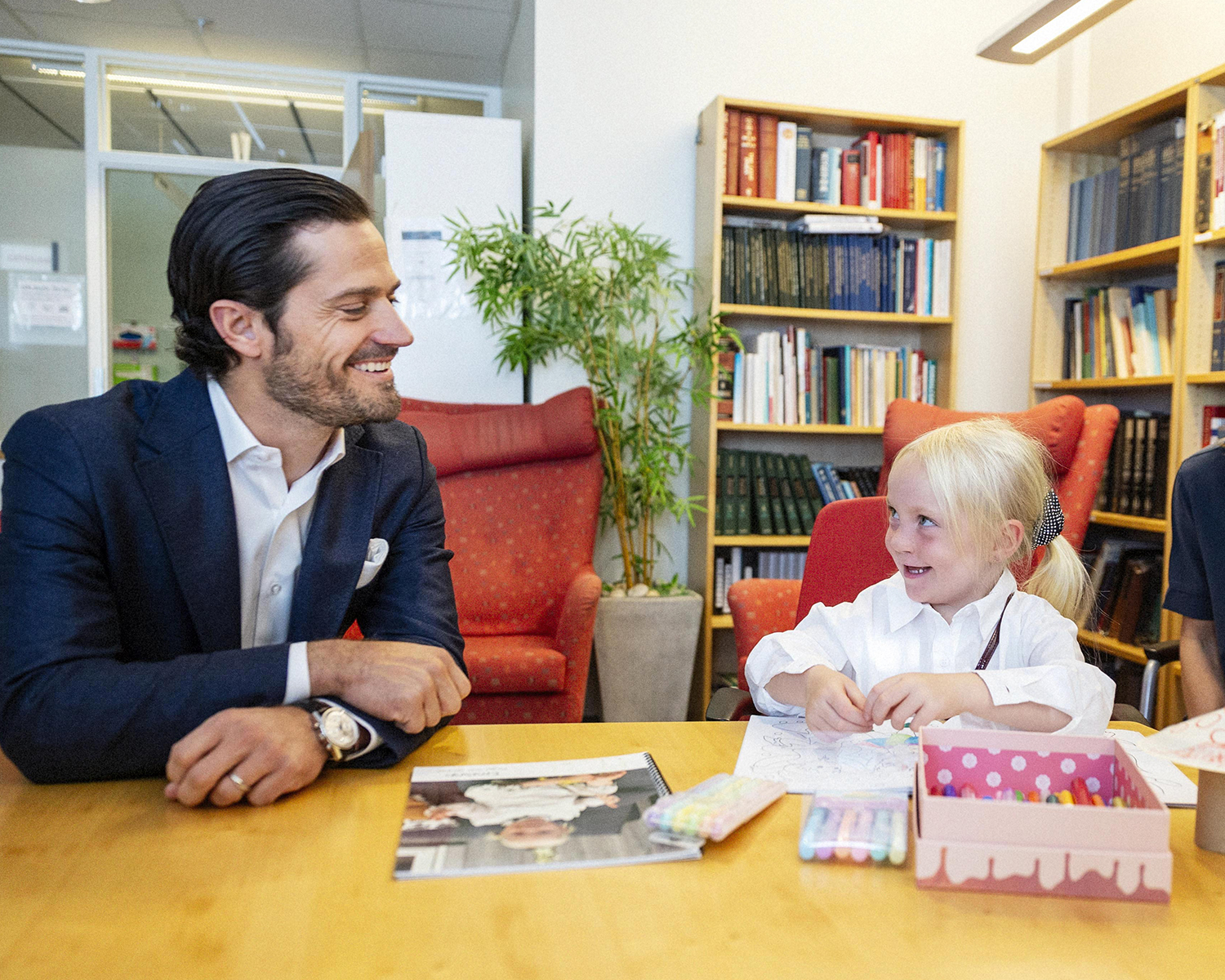 Prins Carl Philip i samtal med 5-årig flicka som pysslar vid ett bord. De sitter nära och tittar på varandra. Båda ser mycket glada ut.