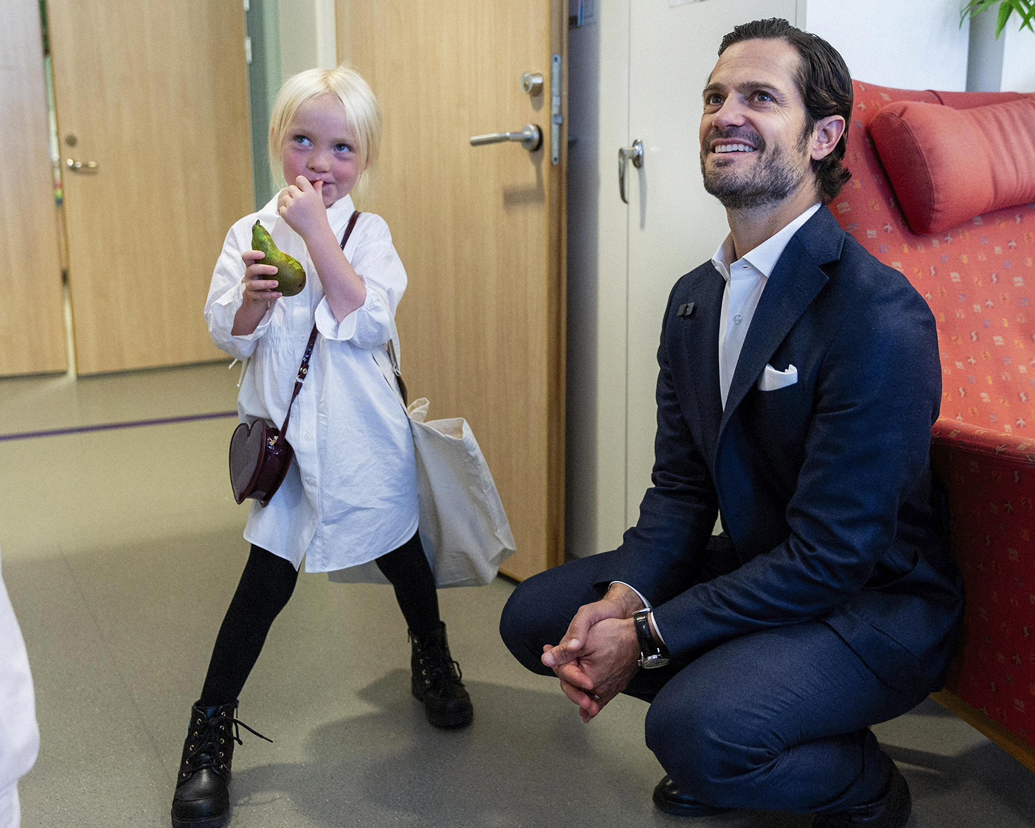 Prins Carl Philip sitter på huk och ler bredvid 5-åriga Emelinn som står bredbent bredvid prinsen med bus i blicken. Båda tittar snett uppåt. Emelinn har två väskor på vardera axeln och ett päron i handen.