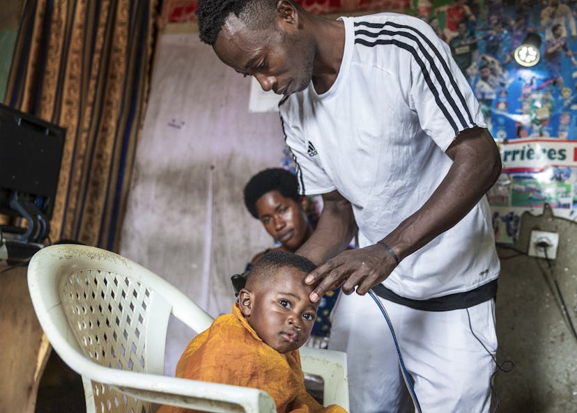 En liten pojke sitter i en plaststol under frisörskynke och blir friserad av sin pappa. Pojken har fått en operation för sin läppspalt.
