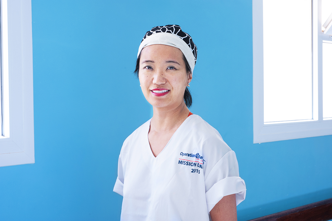 En kvinna med vit vårdklädsel och mönstrad operationsmössa står inomhus mot en blå vägg och ler mot kameran.