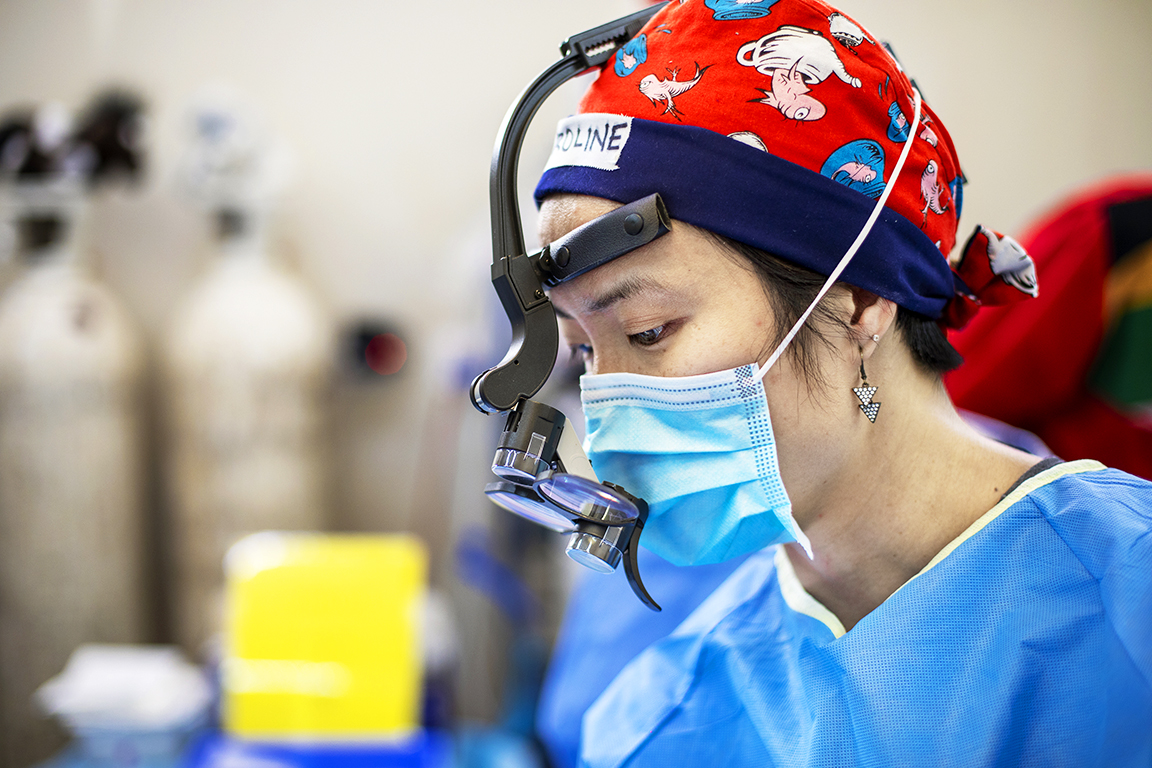 En kirurg med mössa och munskydd använder förstoringsglas vid en operation, syrgastuber och utrustning syns i bakgrunden.