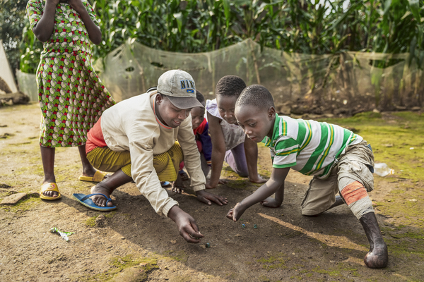 Tre pojkar leker med kulor utomhus. En pojke har kryckor i trä och brännskadade fötter. En pojke har röda shorts och keps.