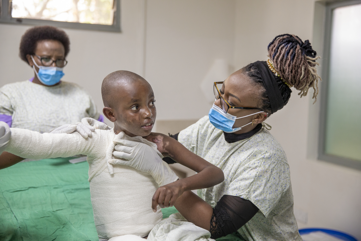 Två vårdpersonal med munskydd och arbetskläder lindar bandage runt ett barns bröstkorg och armar i en vårdmiljö.