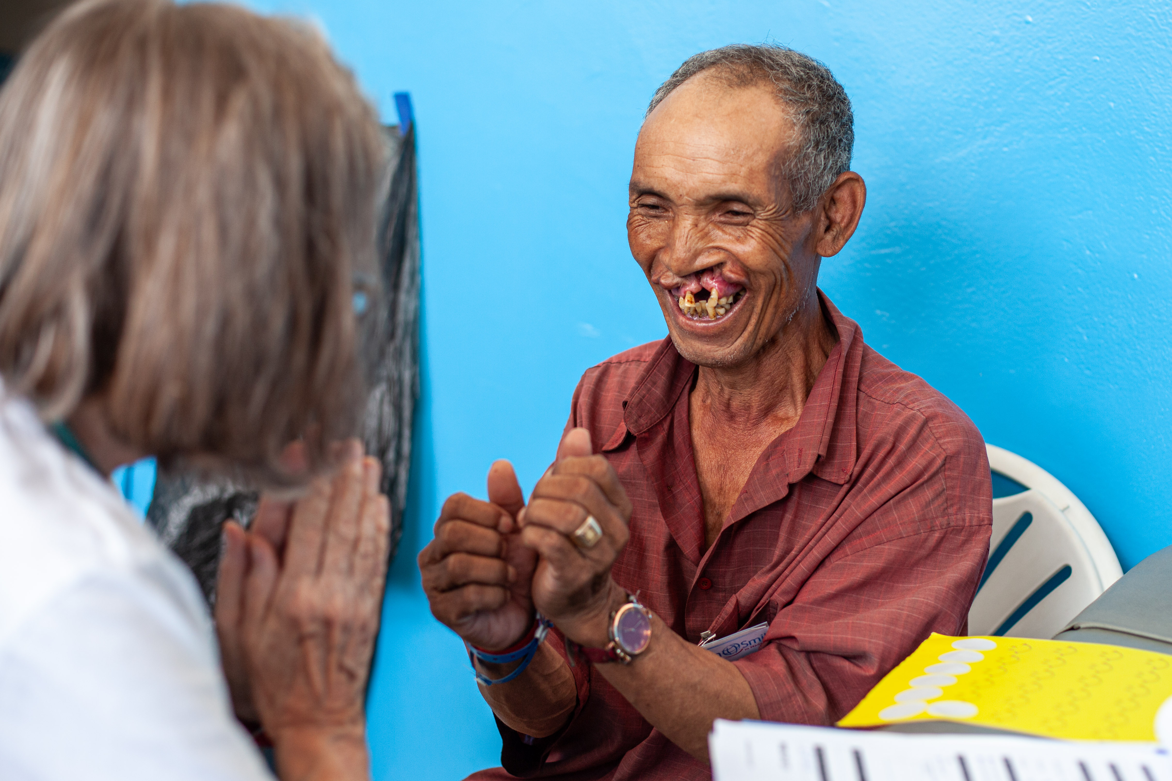 En äldre man med läppspalt och saknade tänder sitter och gestikulerar mot en kvinna med grått hår vid en blå vägg.