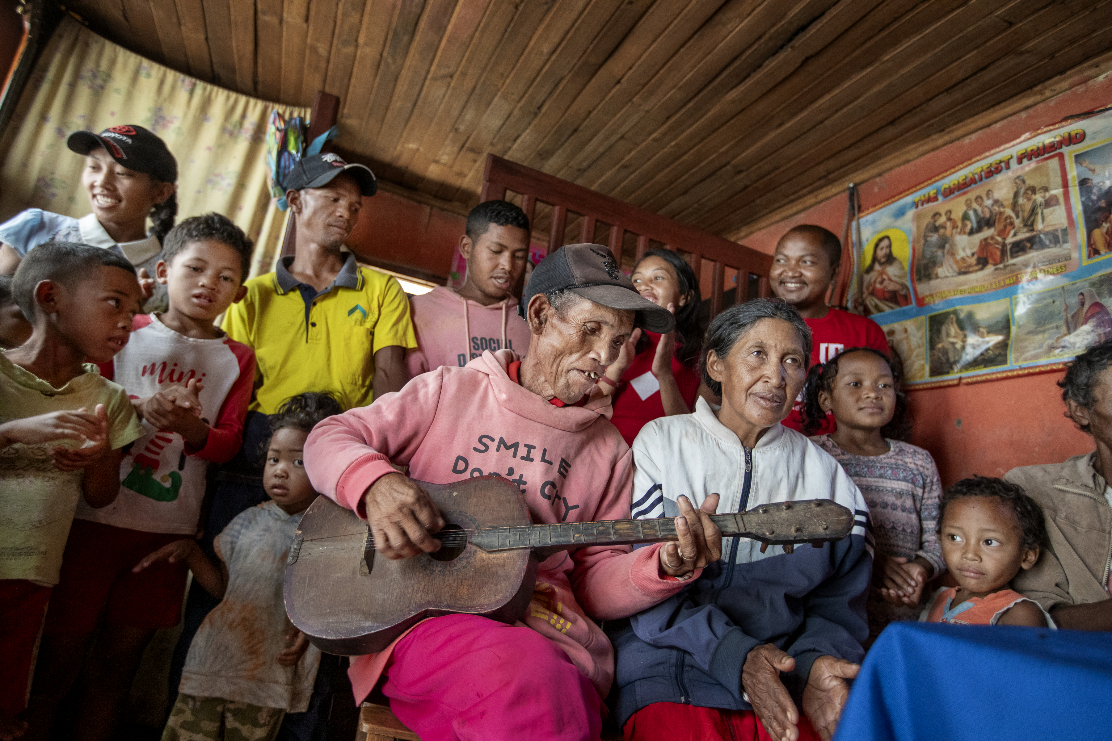 En äldre person spelar gitarr omgiven av barn och vuxna i ett rum med väggar av trä och religiösa affischer.