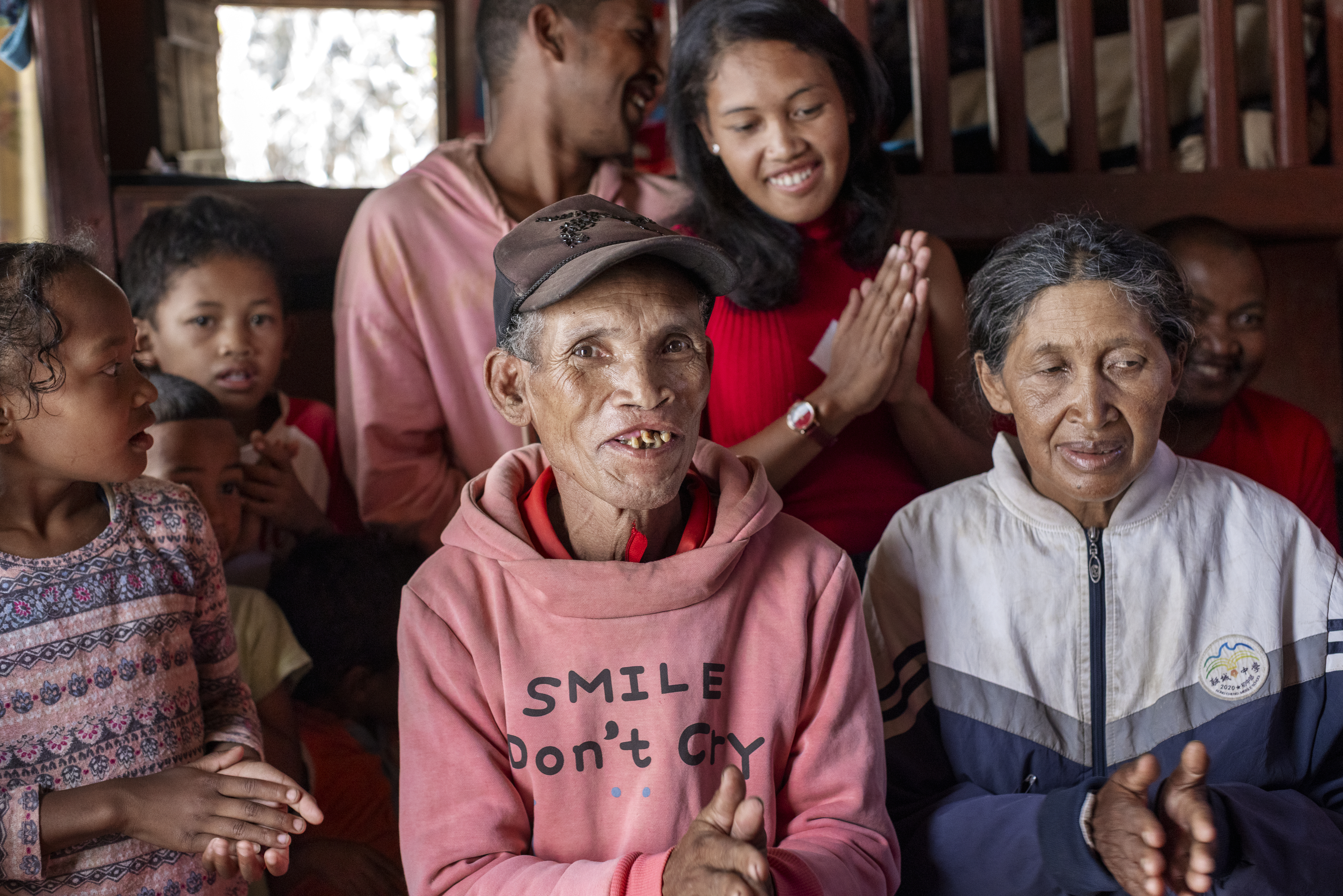 En grupp människor sitter och står inomhus. Några ler och applåderar. Personen längst fram har en rosa hoodie med texten SMILE Dont Cry.