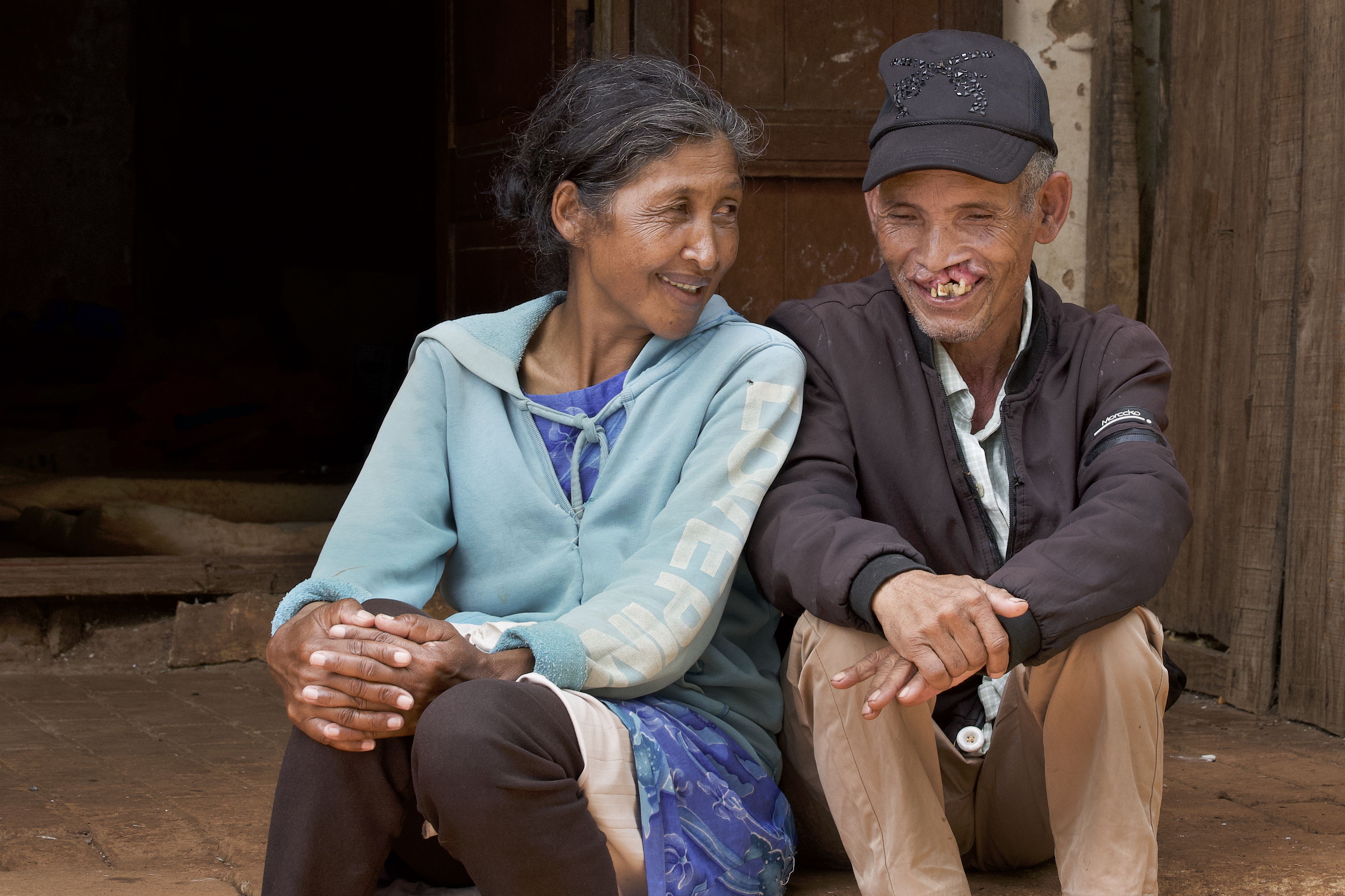 Två vuxna sitter på en trappa och tittar på varandra; kvinnan har en blå huvtröja, mannen har svart keps och jacka.