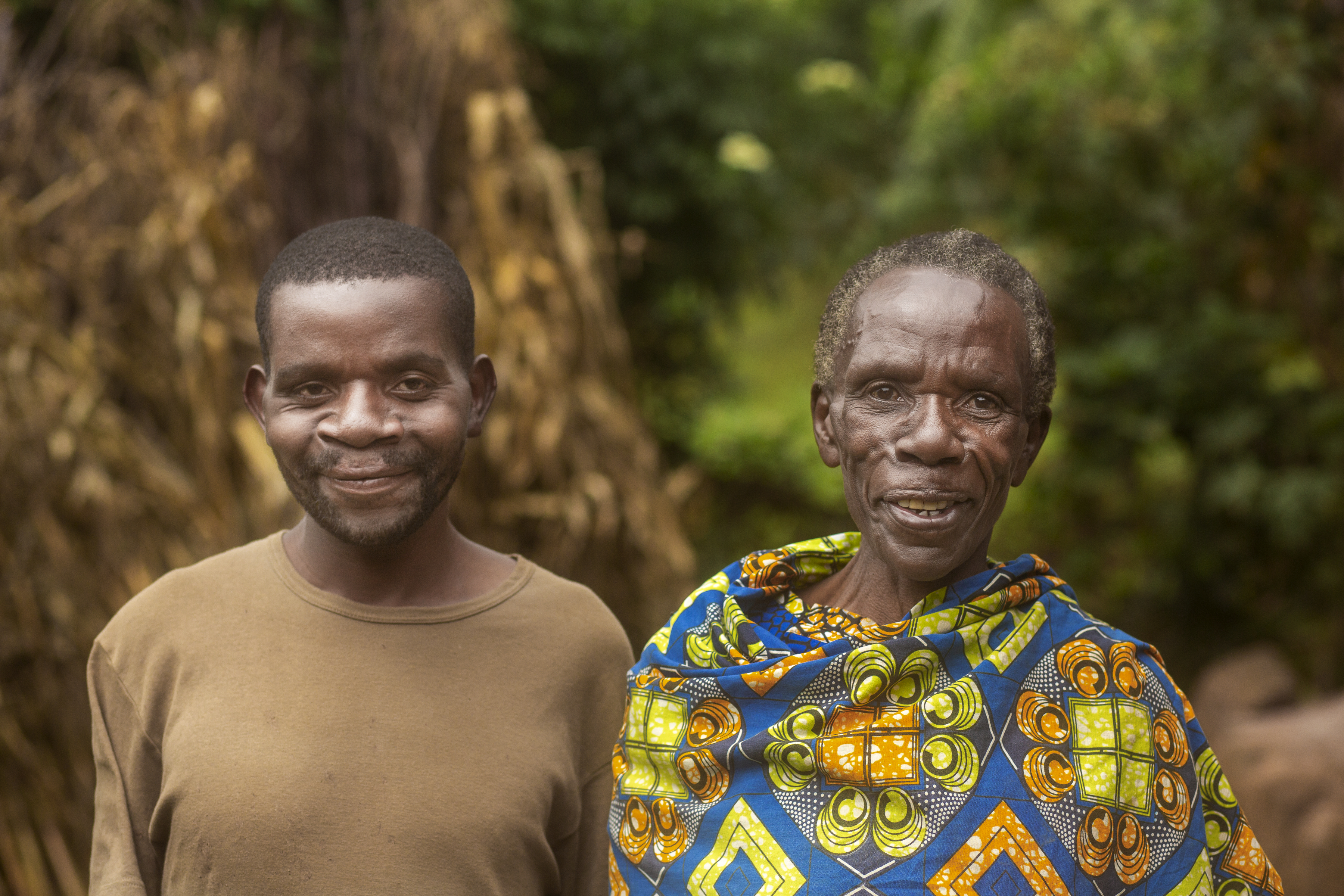 En ung man bredvid en äldre man ser in i kameran och ler. Den äldre mannen har en färgglad sarong över sina axlar. Den yngre mannen har en mörk-beige tröja.
