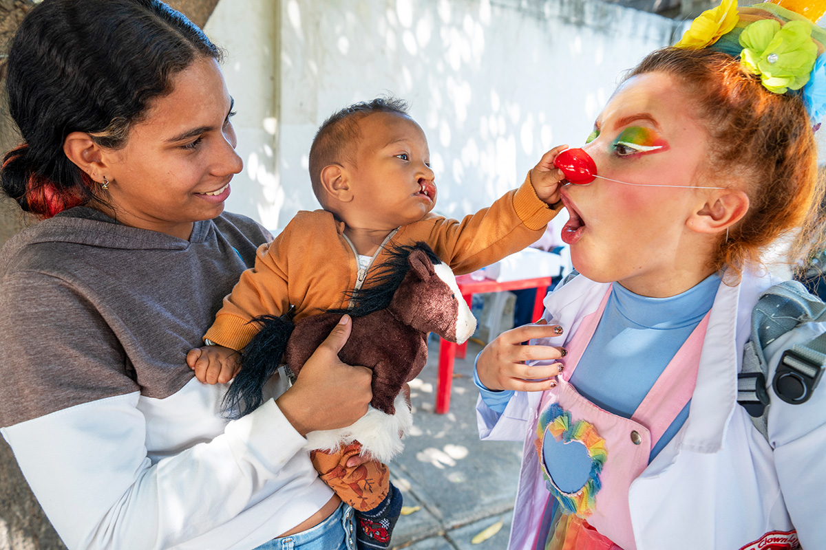 En kvinna håller i en bebis som rör vid den röda näsan på en person utklädd till clown.