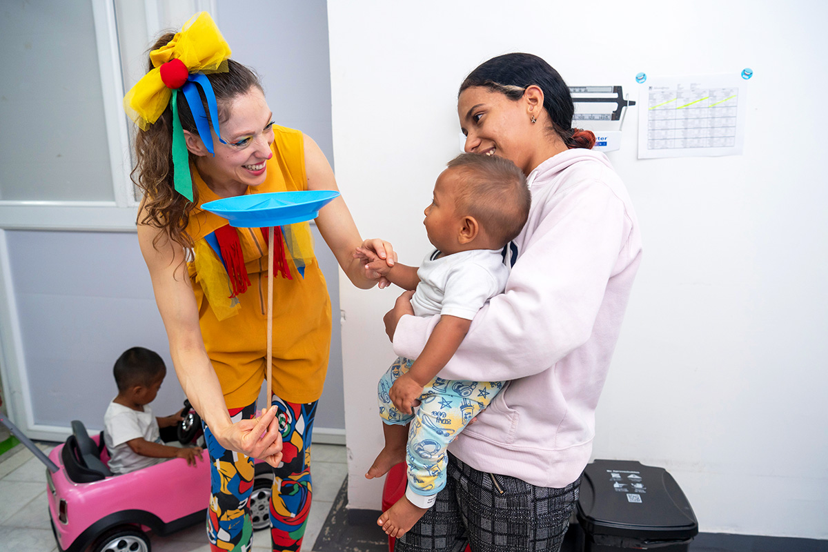 En kvinna utklädd till clown snurrar en tallrik på en pinne för ett spädbarn i famnen på en vuxen, med ett barn i bakgrunden.