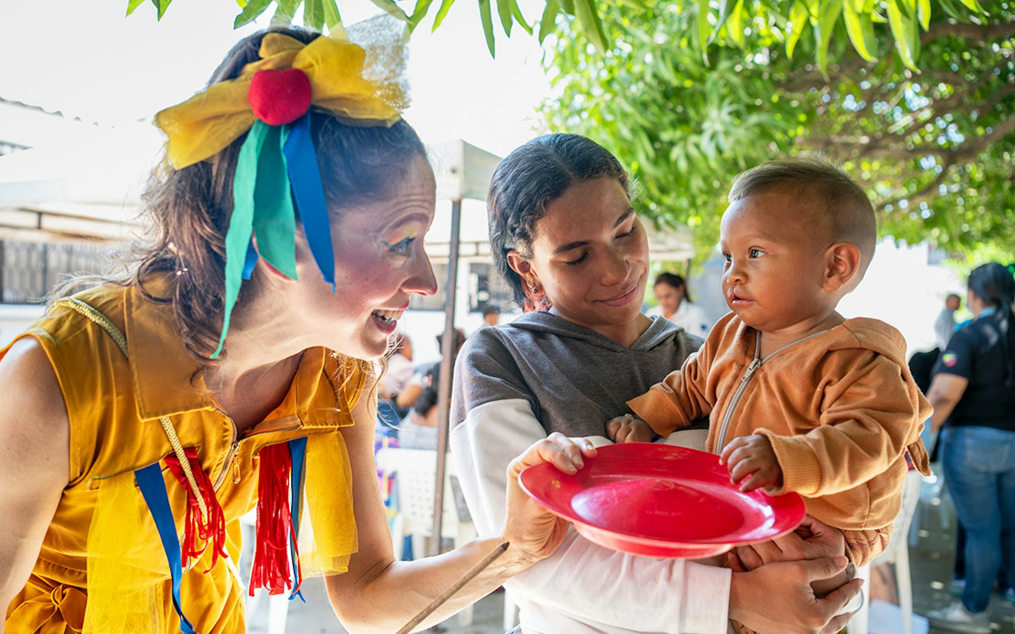 En kvinna i dräkt pratar med en mamma som håller ett barn och en röd tallrik vid ett utomhusevenemang.