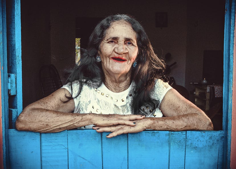 Dona Maria, en 67-årig kvinna från Brasilien står lutat mot en blå halvdörr. Hon har rött läppstift och ler.
