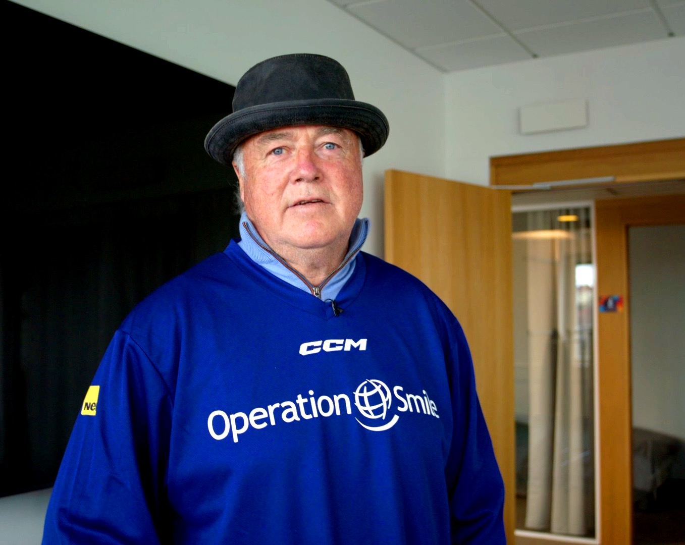 En äldre man med svart hatt och blå Operation Smile-tröja står inomhus nära en trädörr och ett glasat kontorsutrymme.