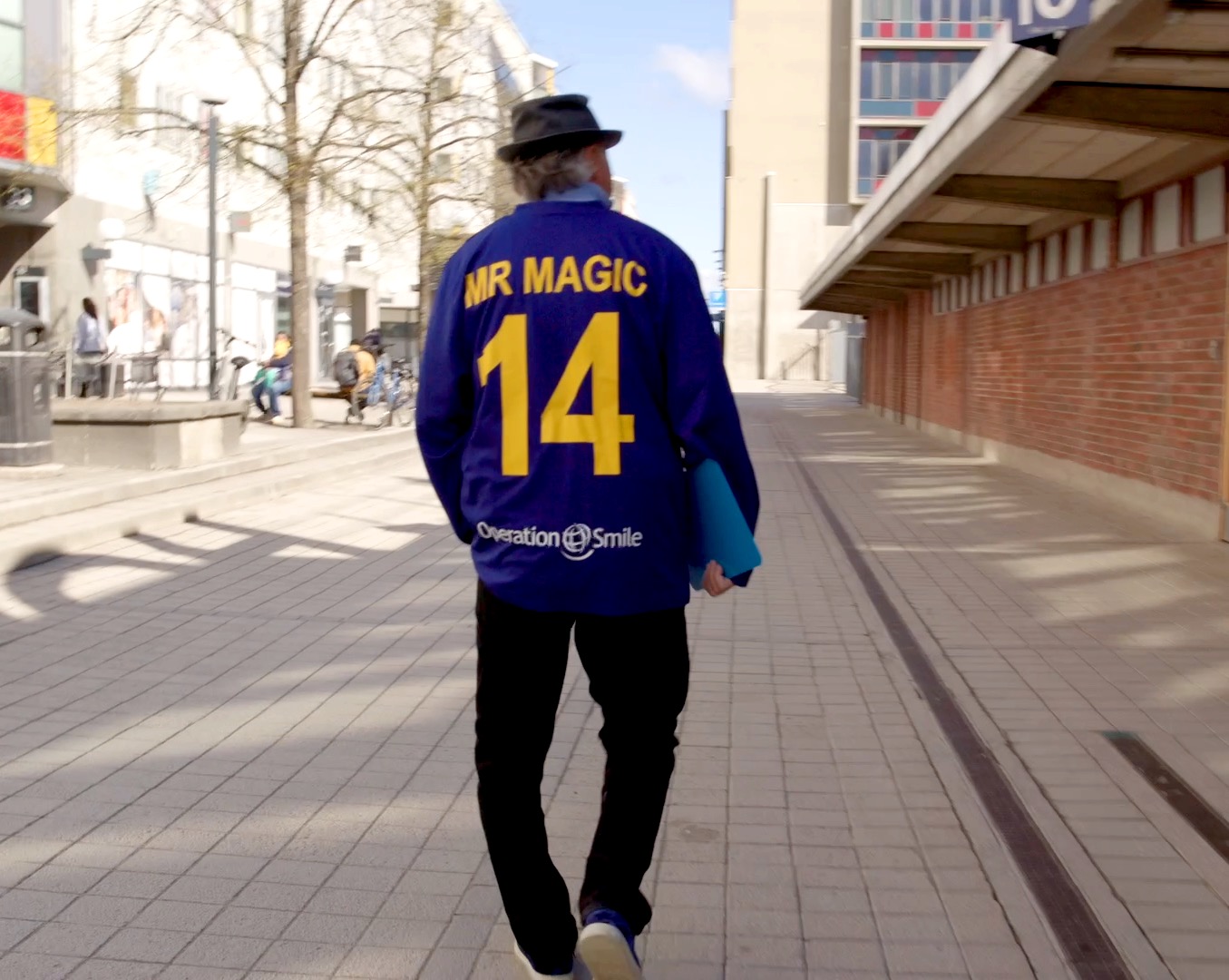 En person med blå jacka där det står MR MAGIC 14 och Operation Smile på ryggen går på en trottoar och håller en blå mapp.