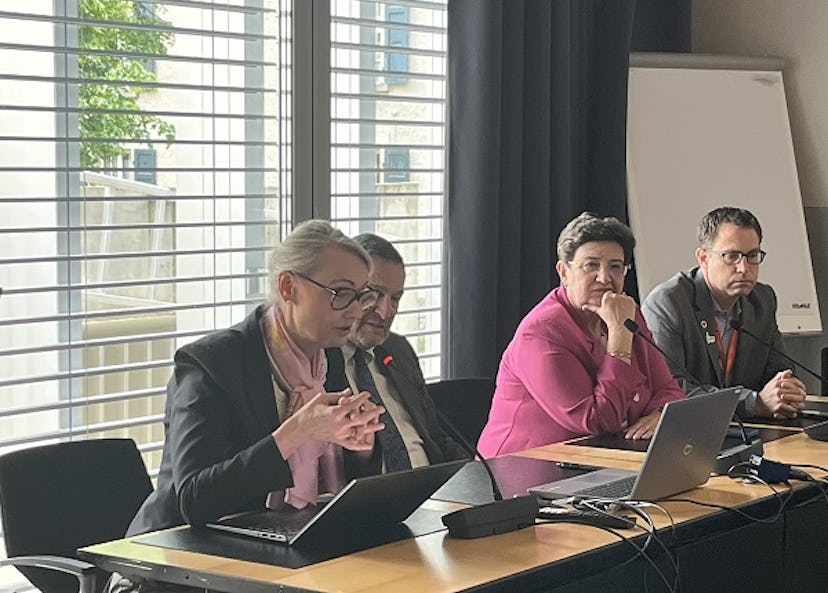 Fyra personer sitter vid ett konferensbord med bärbara datorer under en paneldiskussion. En kvinna talar, de andra lyssnar. Ett tomt blädderblock syns i bakgrunden.