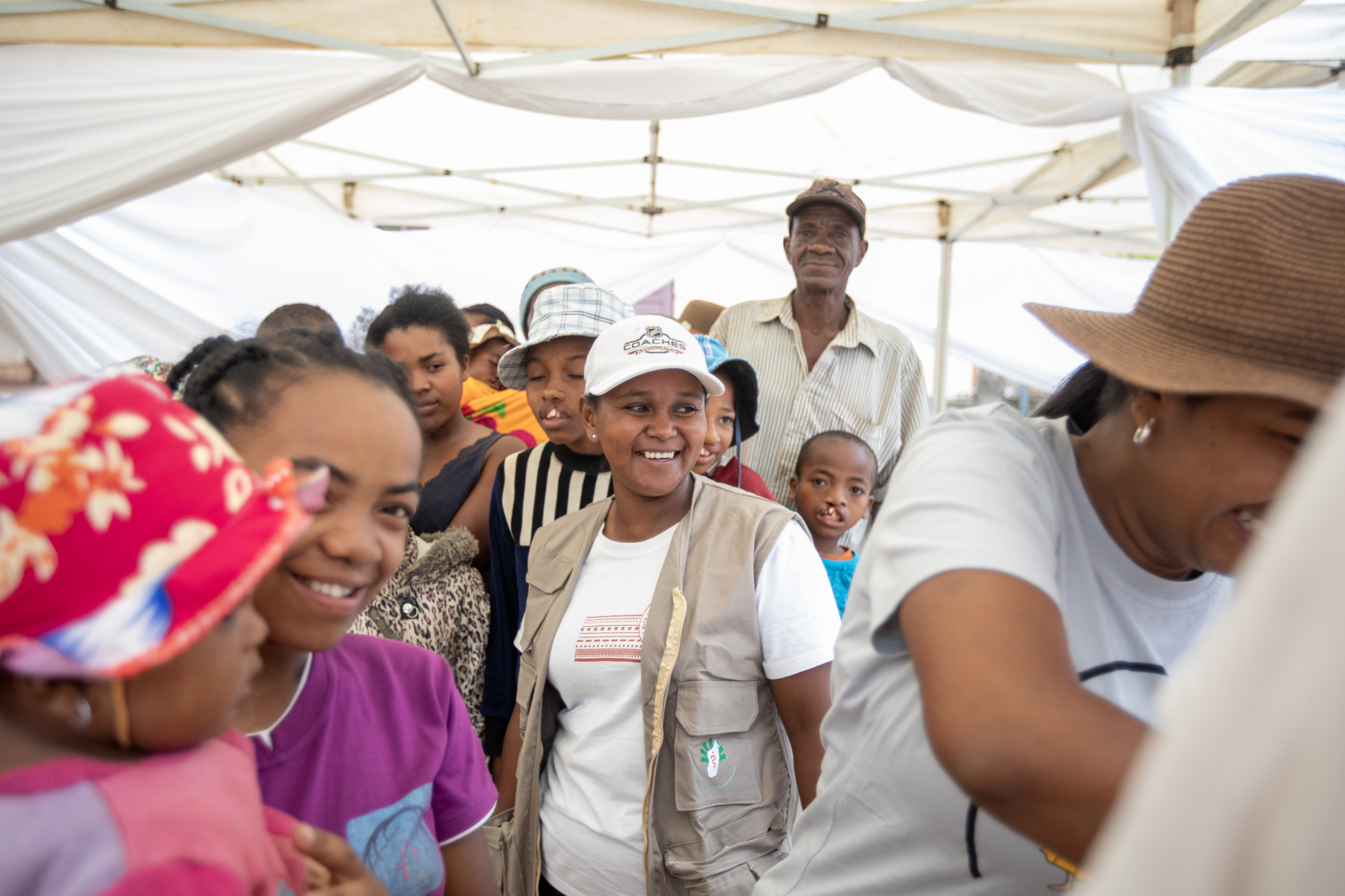 En grupp människor, både barn och vuxna, står samlade under ett tält. En kvinna i mitten har vit keps och beige väst.