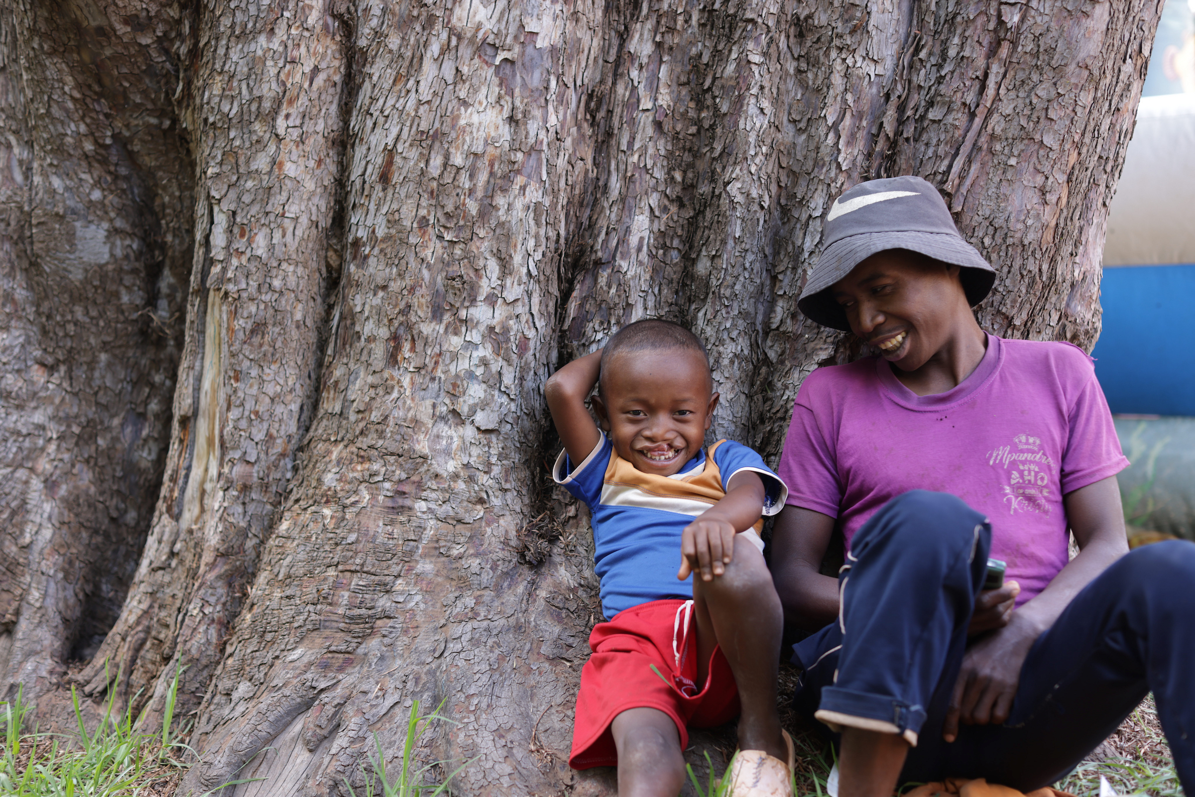 En liten pojke med läppspalt sitter lutad mot en kraftig trädstam med sin pappa bredvid sig.