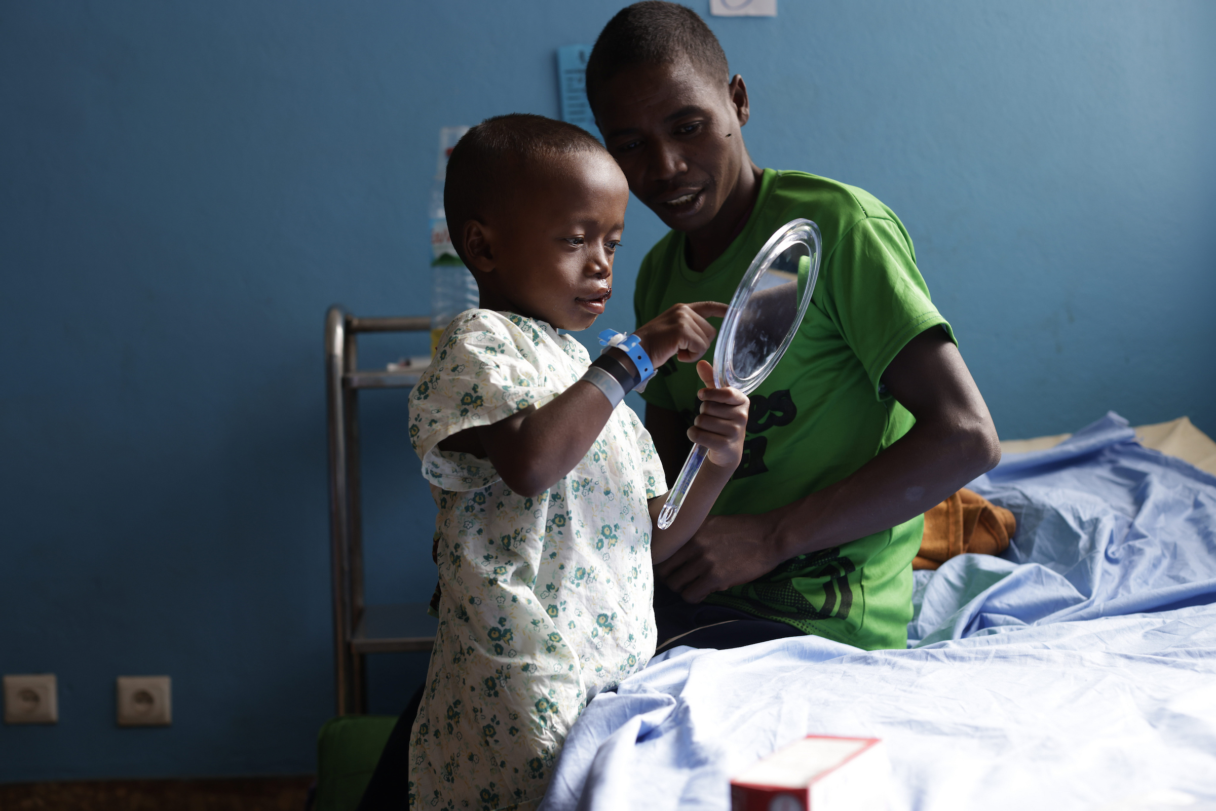 En liten pojke i sjukshuslinne håller i en handspegel och pekar på sin egen spegelbild. Han ser glad och nyfiken ut efter att ha fått en spaltoperation. Hans pappa sitter i grön t-shirt bredvid honom på sjukhussängen.