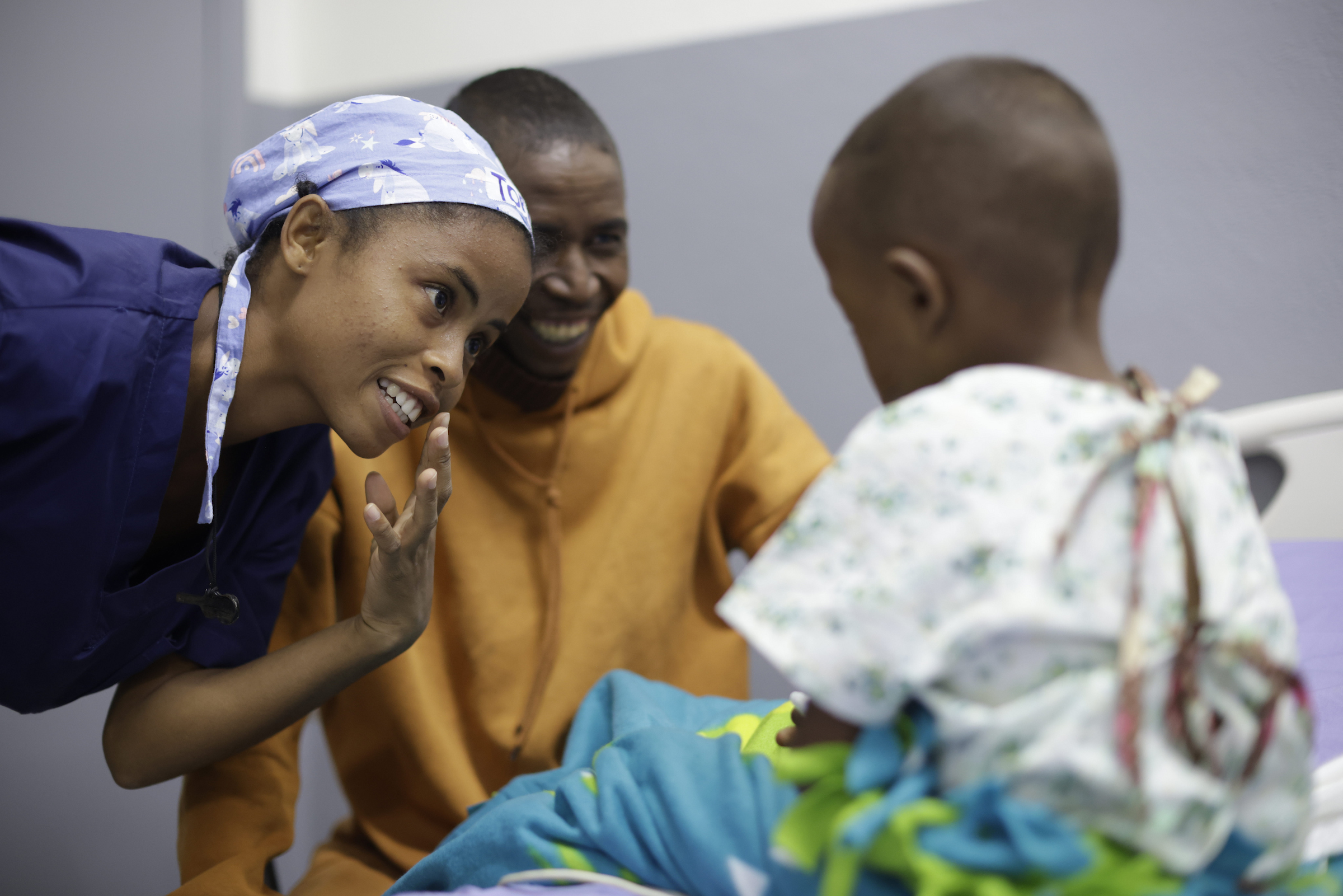 En sjuksköterska lutar sig ned intill en liten pojke som vaknat upp från sin läppspalt-operation. Pappan sitter glad och lättad bredvid.