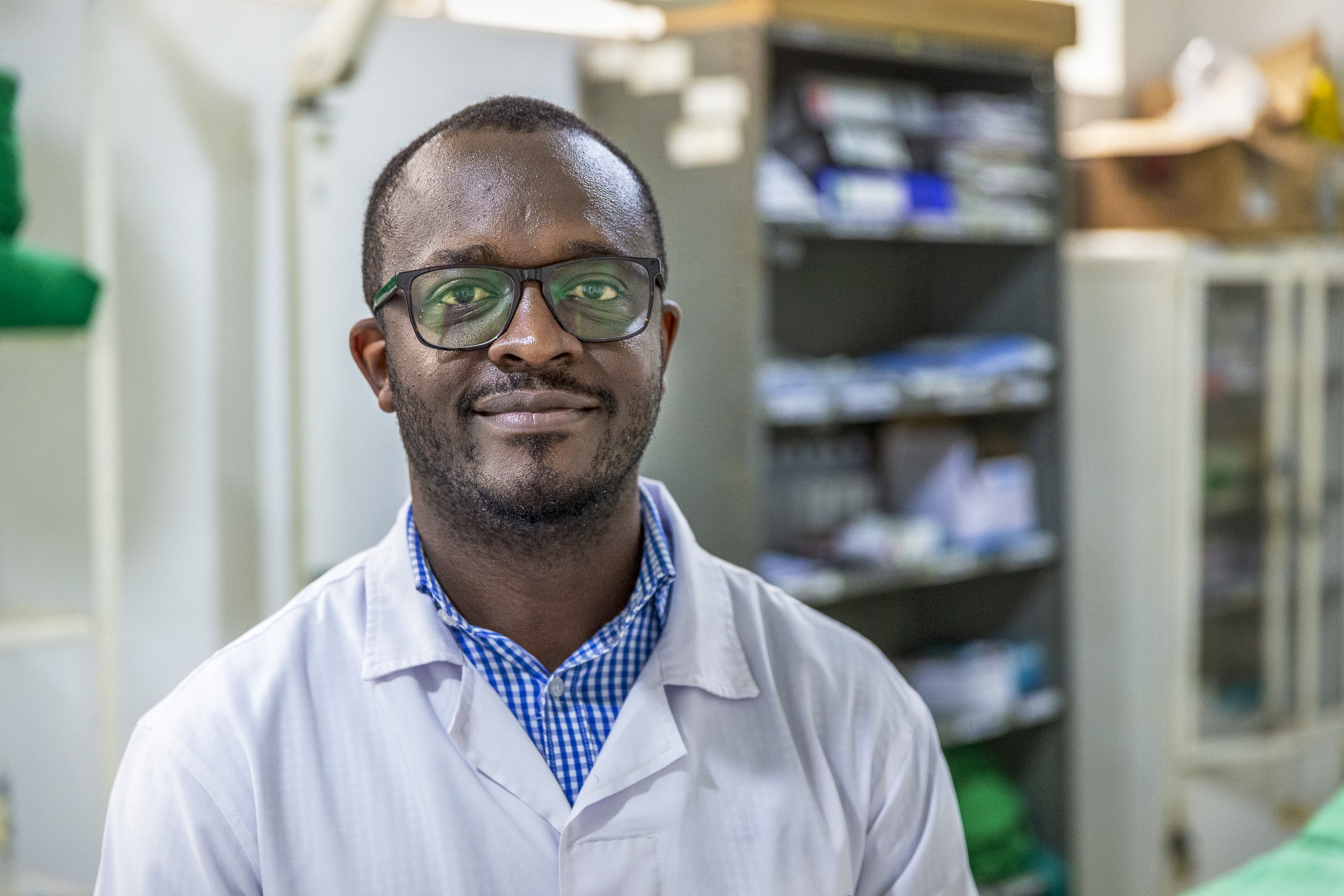 En man med glasögon och vit labbrock står i ett rum med hyllor som har medicinska förnödenheter.