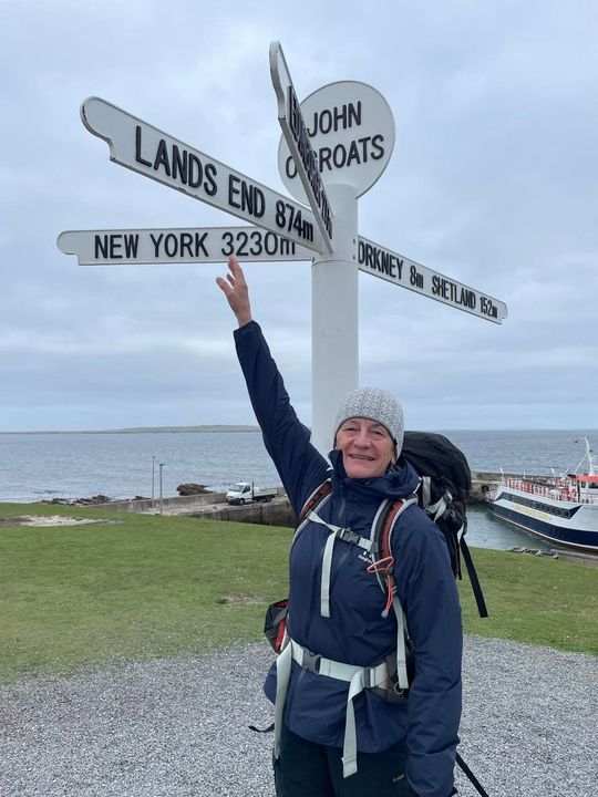 En person i friluftskläder står vid en vägskylt med destinationer som Land's End och New York, med en färja och hav i bakgrunden.