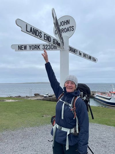 En person i friluftskläder står vid en vägskylt med destinationer som Land's End och New York, med en färja och hav i bakgrunden.