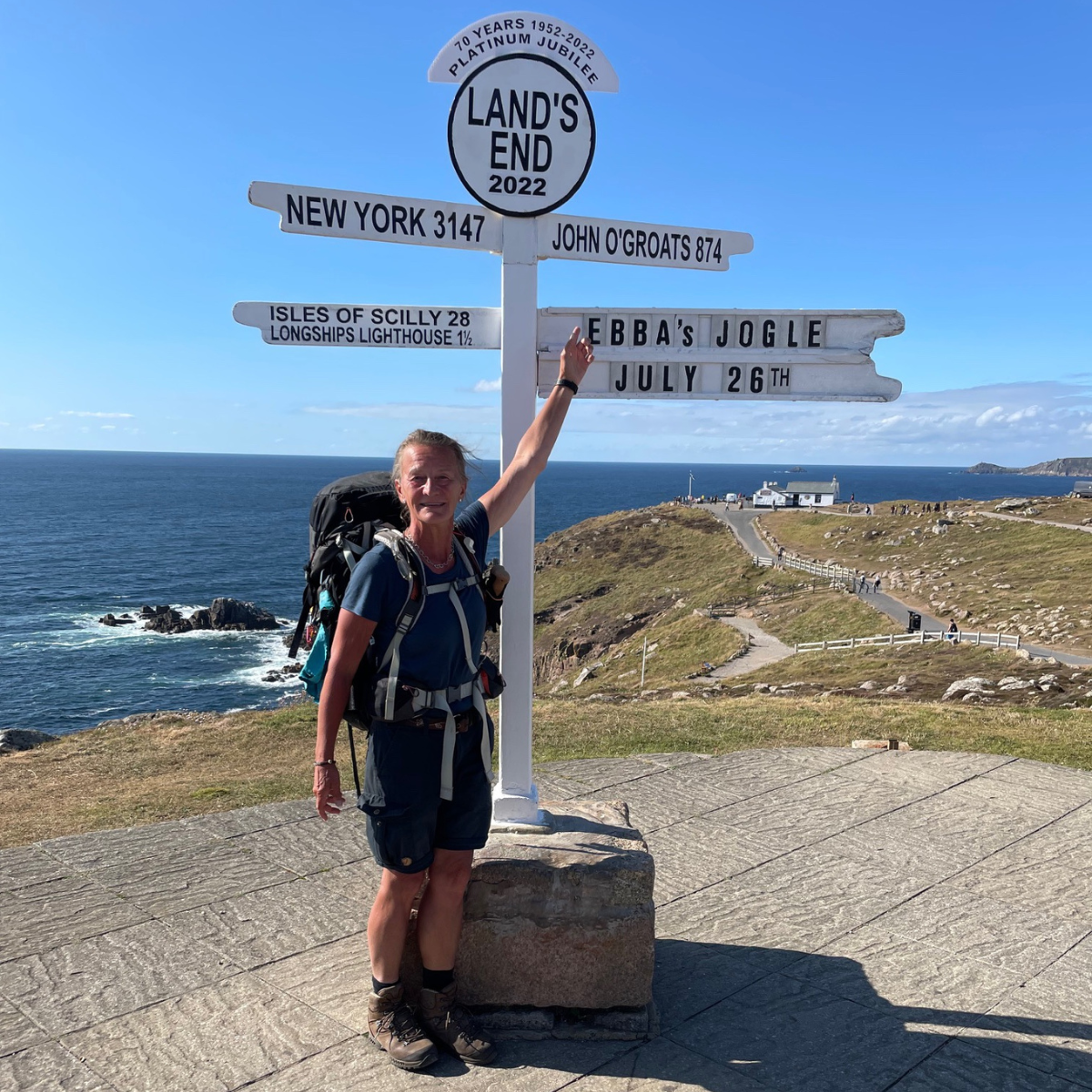 En person med stor ryggsäck står vid skylten Land's End och pekar på en tavla med texten "EBBAS JOGLE JULY 26TH", kusten syns bakom.