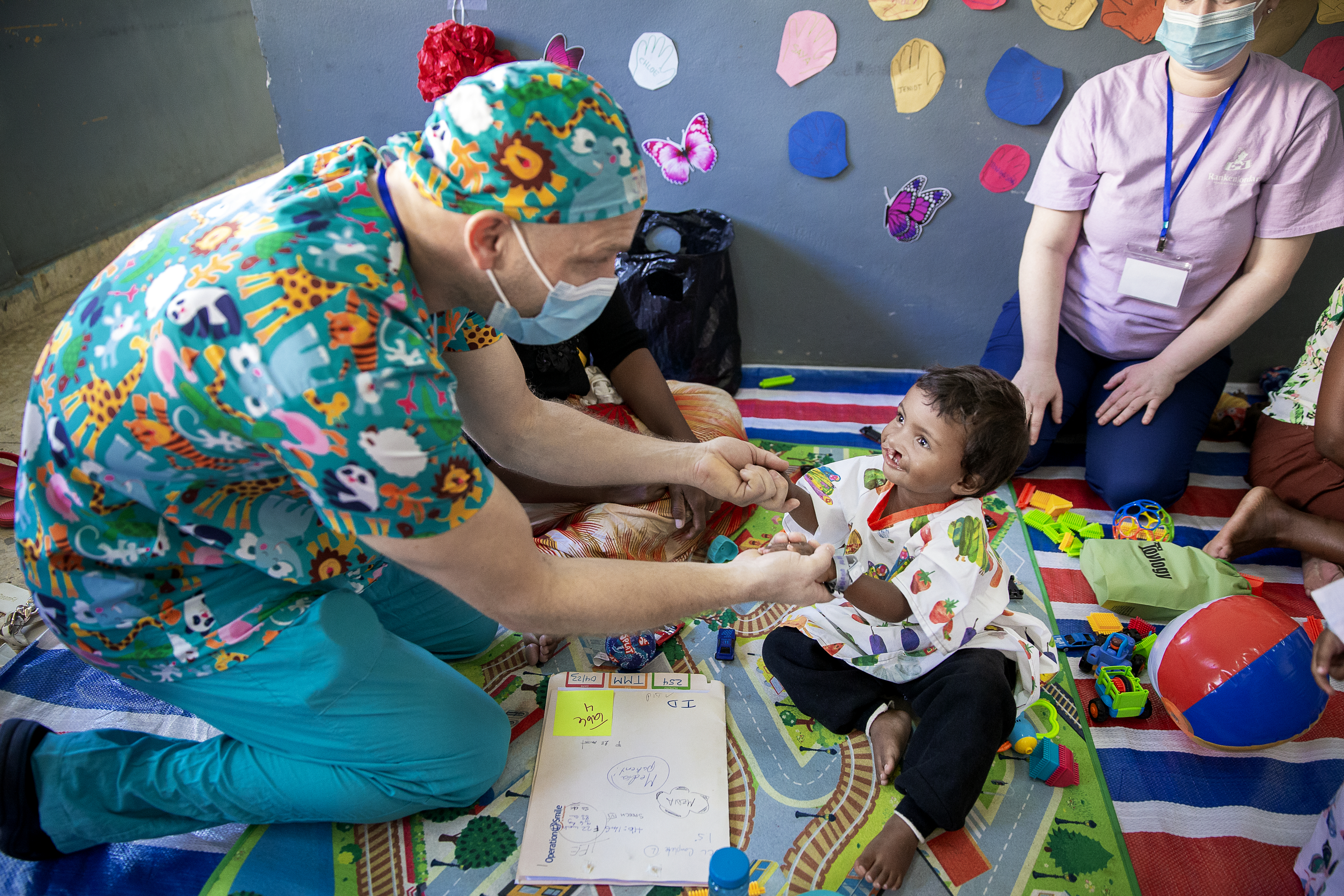 En vårdarbetare i vårdkläder pratar med ett barn som sitter på en matta med leksaker, medan andra ser på i ett lekrum.