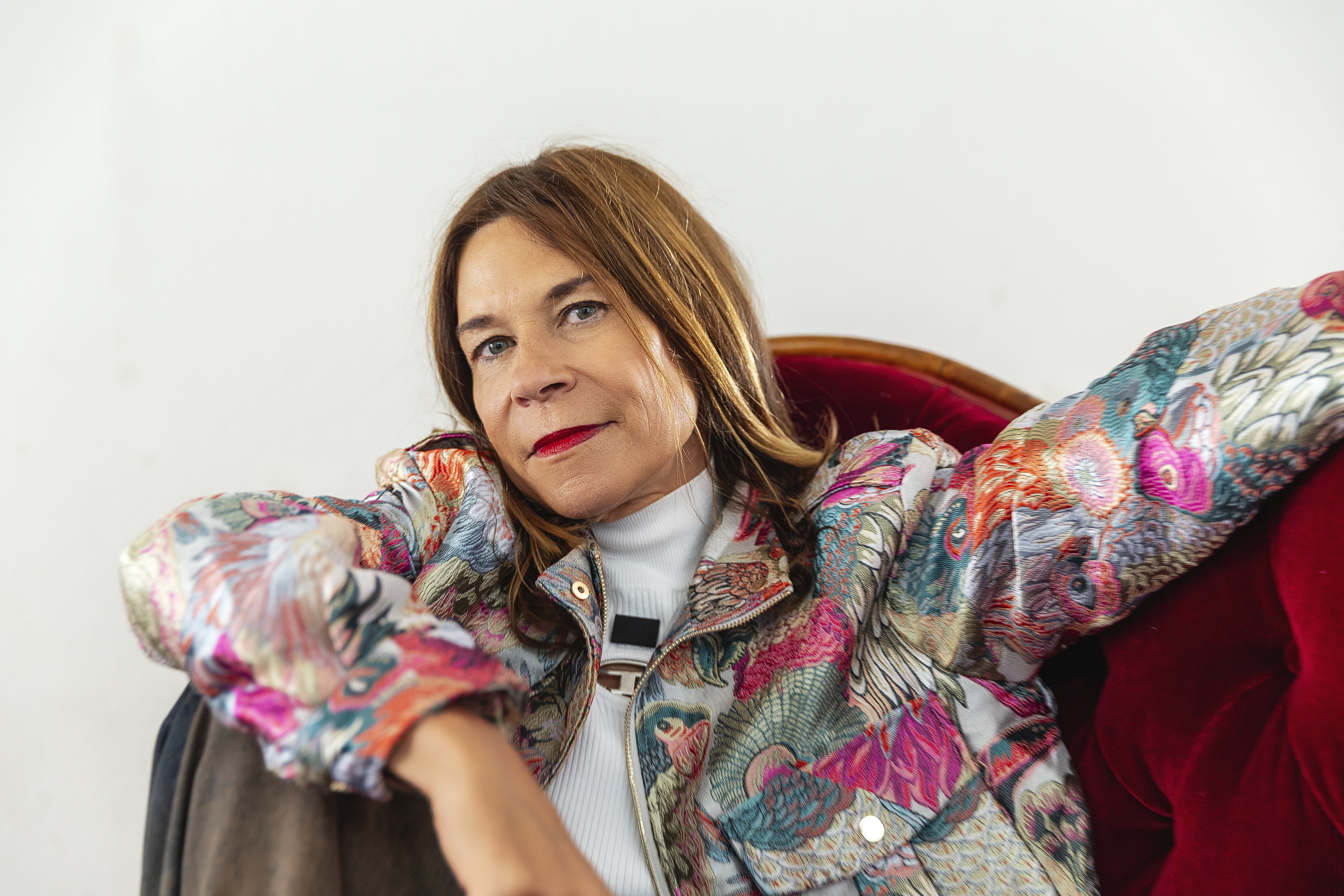 En kvinna med rakt brunt hår sitter på en röd stol, har på sig en mönstrad jacka och en vit polotröja och tittar in i kameran.