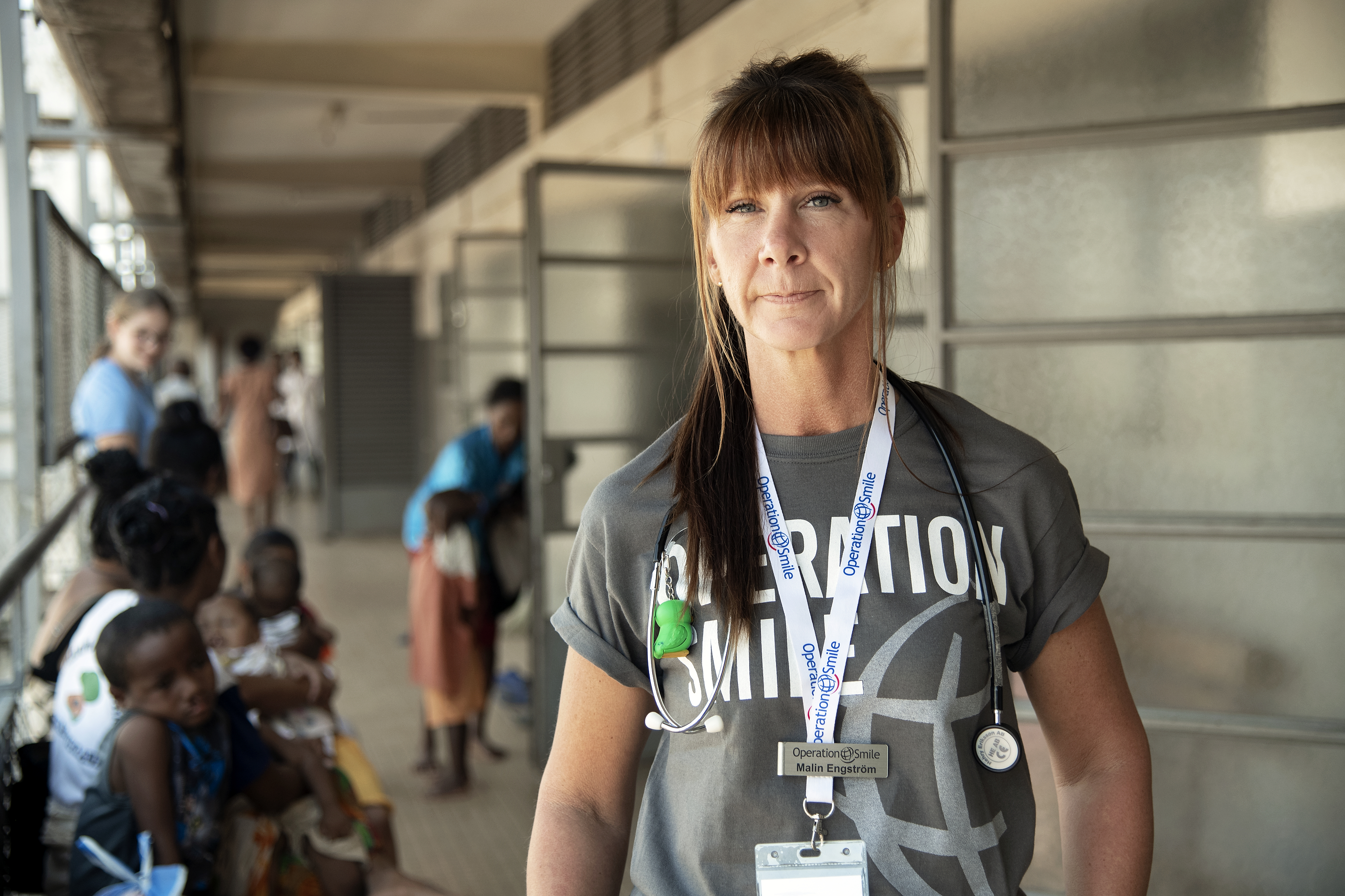 En kvinnlig vårdarbetare i en "Operation Smile"-tröja står i en korridor; familjer med barn väntar på bänkar längs väggen i bakgrunden.