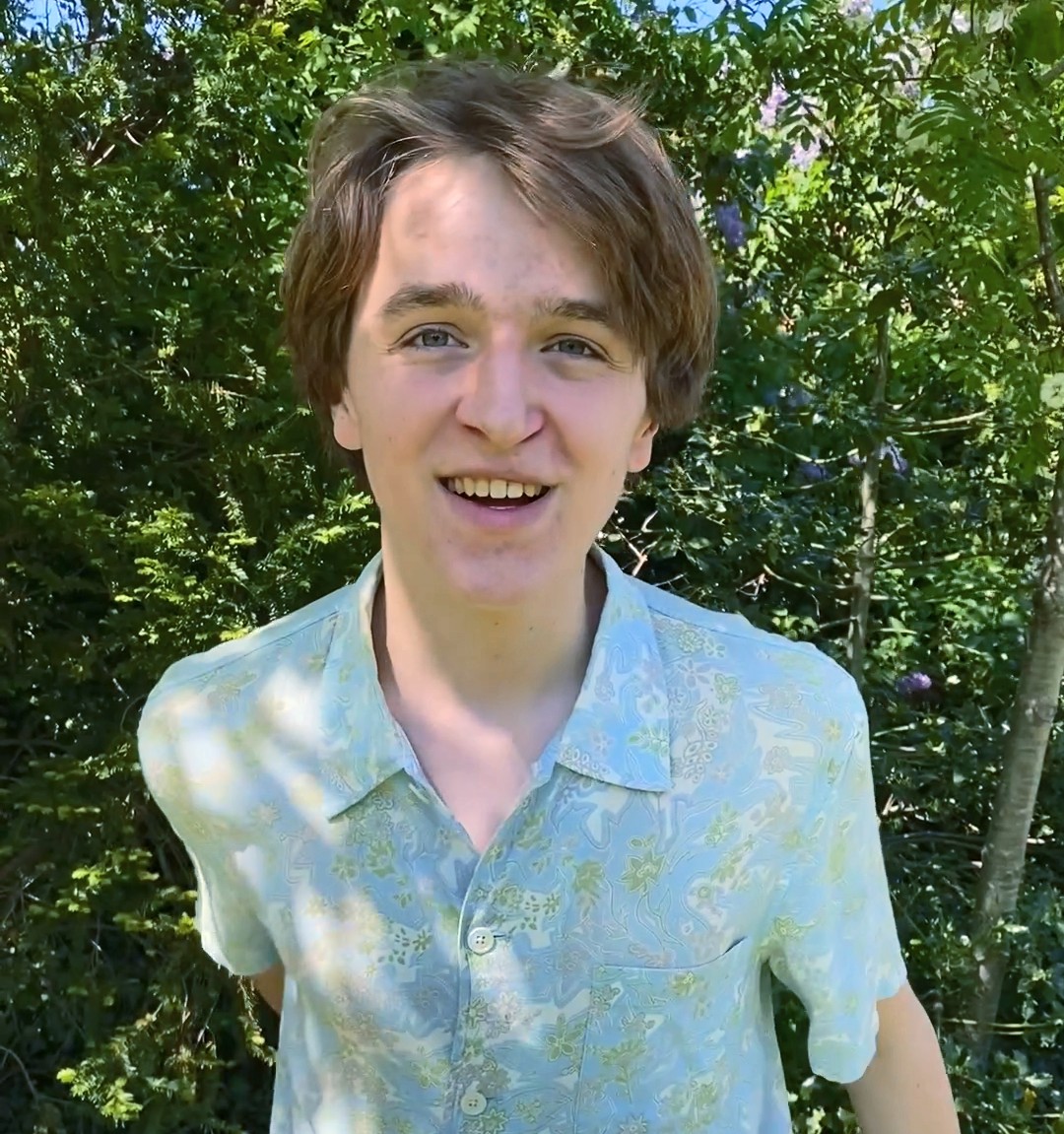En ung person med ljusbrunt hår och en ljusmönstrad skjorta står utomhus framför grönska och ler mot kameran.