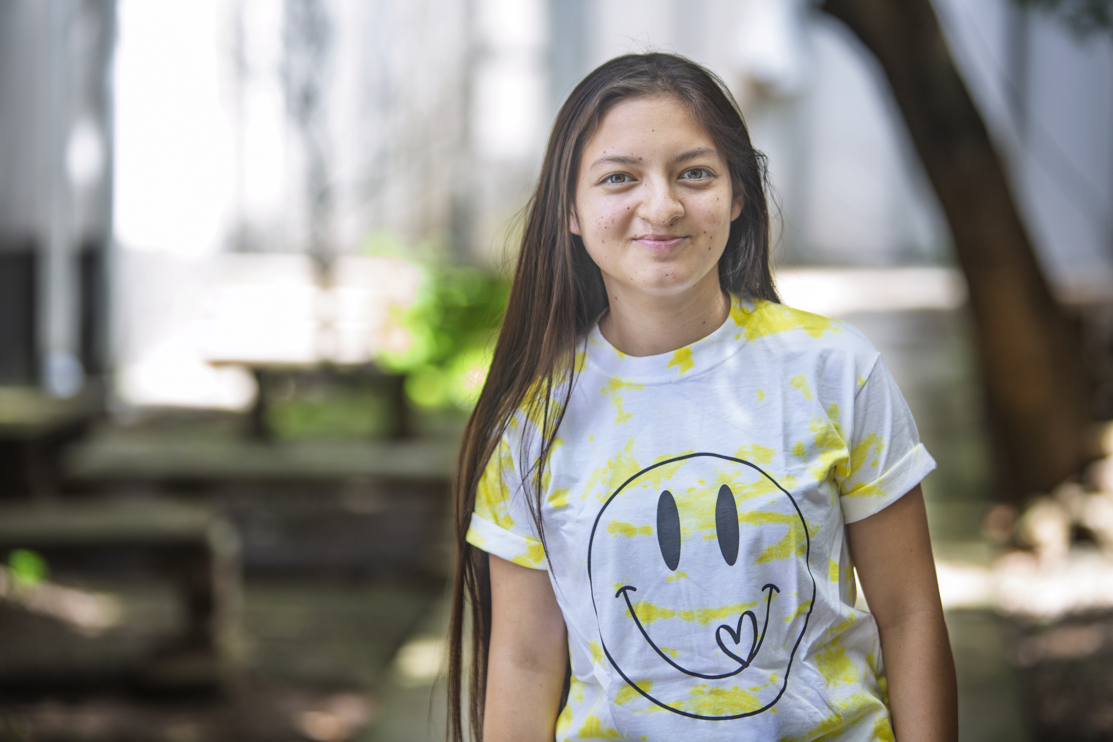 En ung kvinna med långt brunt hår står utomhus och har på sig en vit och gul tröja med ett stort smileymotiv. Träd och bänkar syns i bakgrunden.