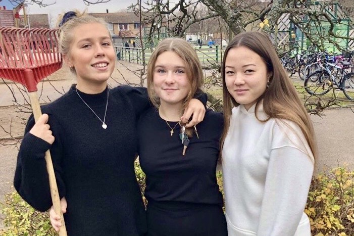 Tre unga kvinnor står utomhus, en håller i en röd kratta, poserar och ler mot kameran med byggnader och cyklar i bakgrunden.