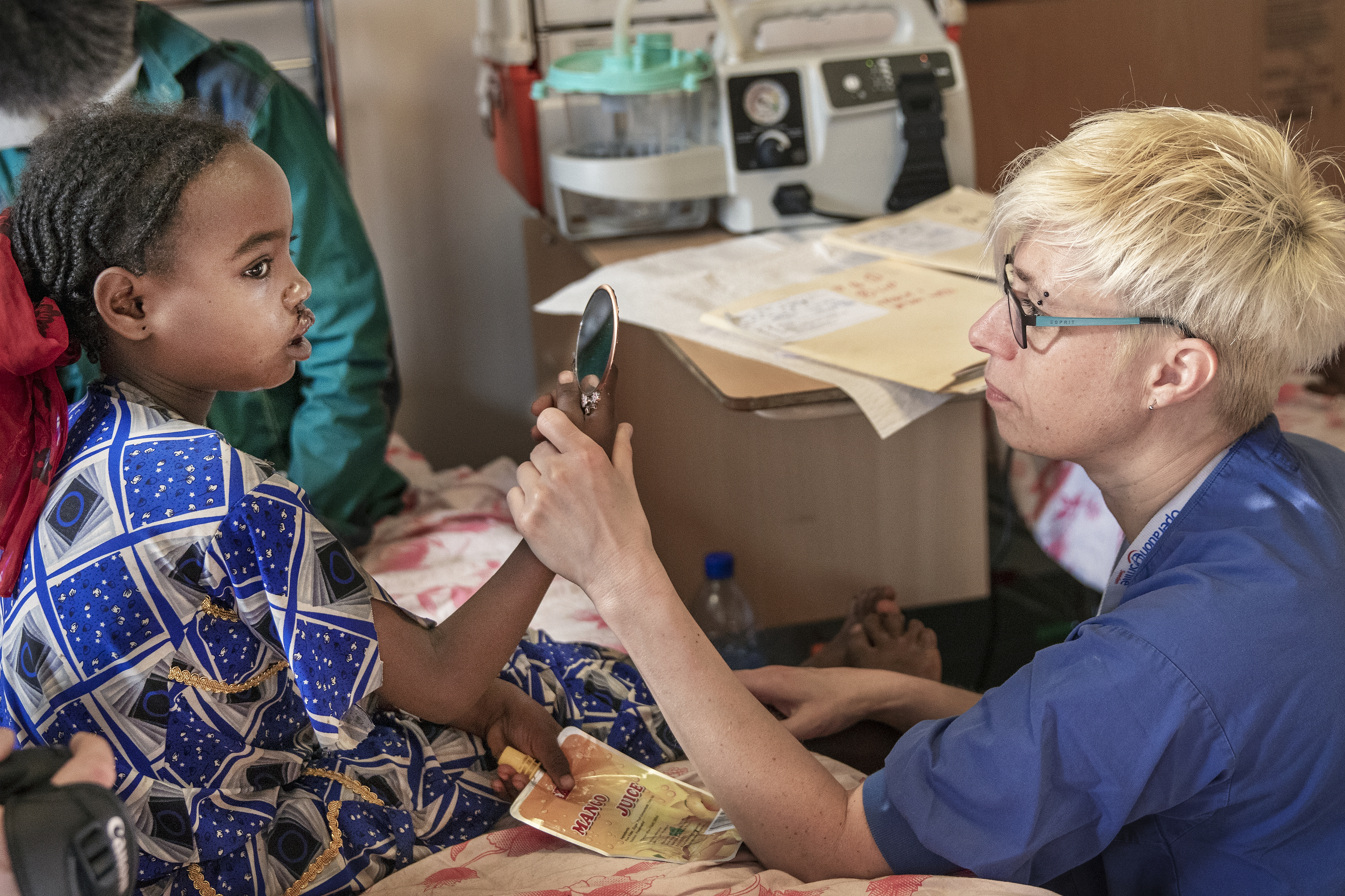 En vårdarbetare håller upp en liten spegel åt en ung flicka som sitter på en sjukhussäng, med medicinsk utrustning och papper i bakgrunden.