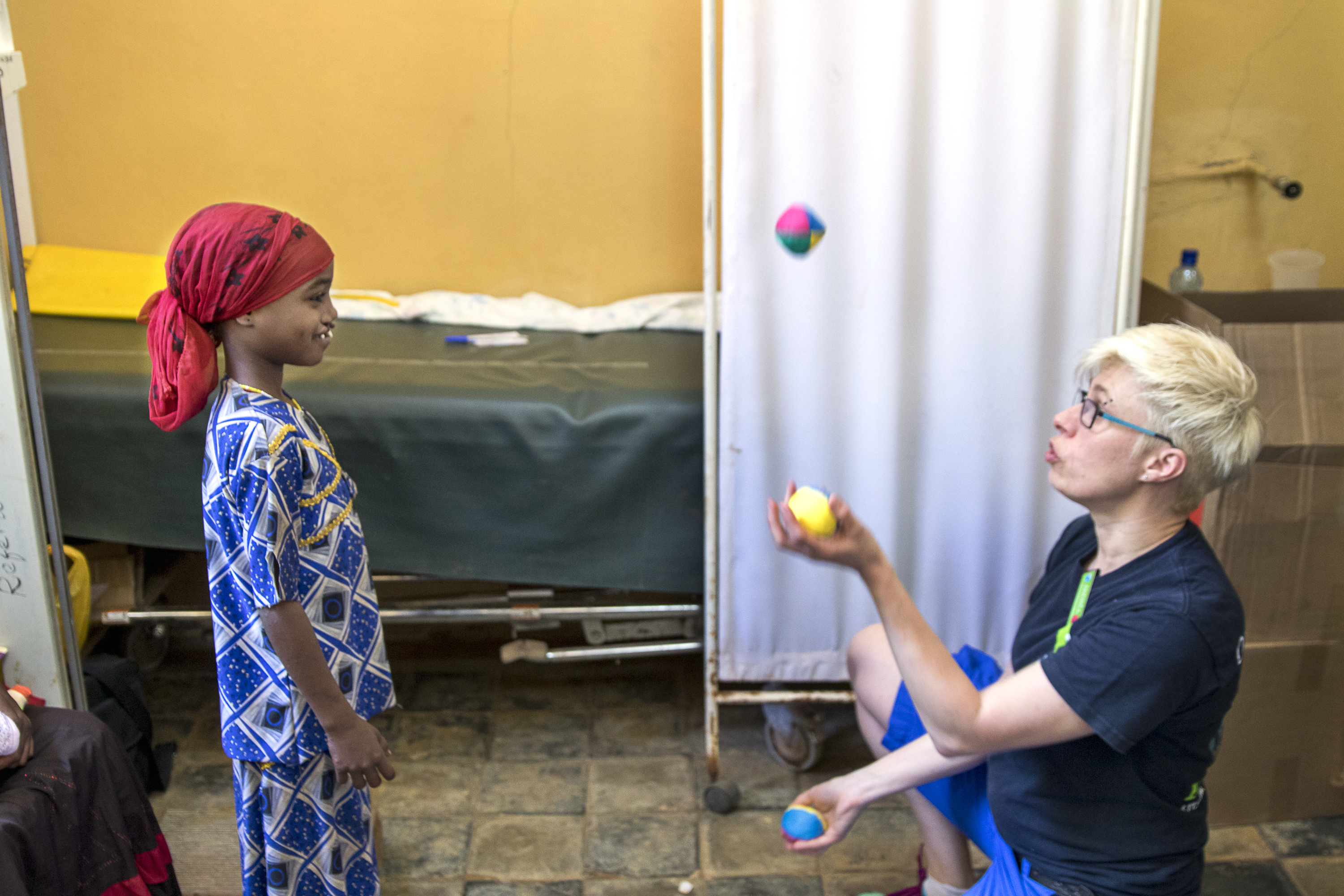 En vuxen jonglerar med bollar medan ett barn i mönstrade kläder och röd huvudduk tittar på bredvid en sjukhussäng och gardin.