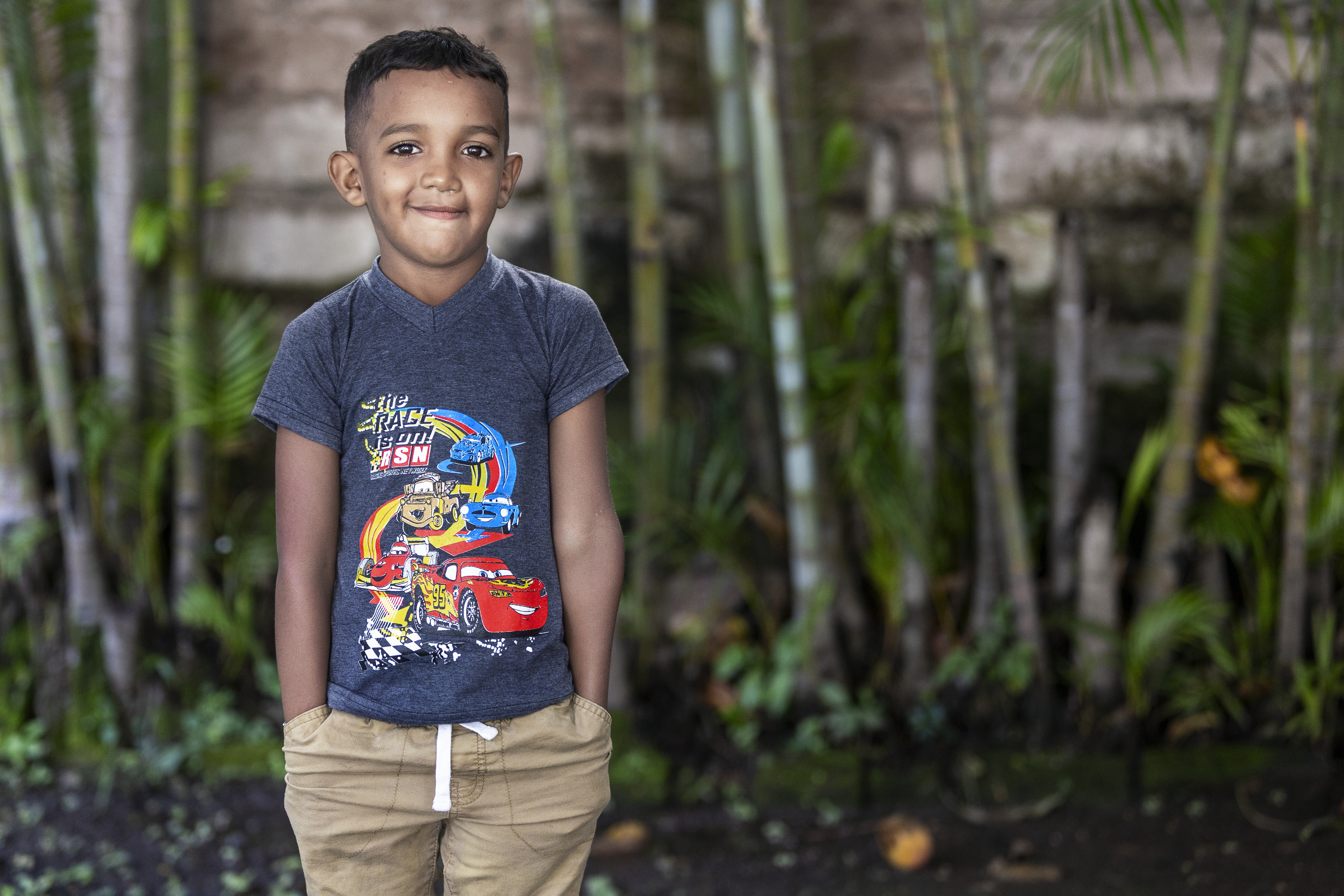 En pojke står utomhus med händerna i fickorna, har på sig en t-shirt med en tecknad figur och shorts, framför växter och en stenmur.