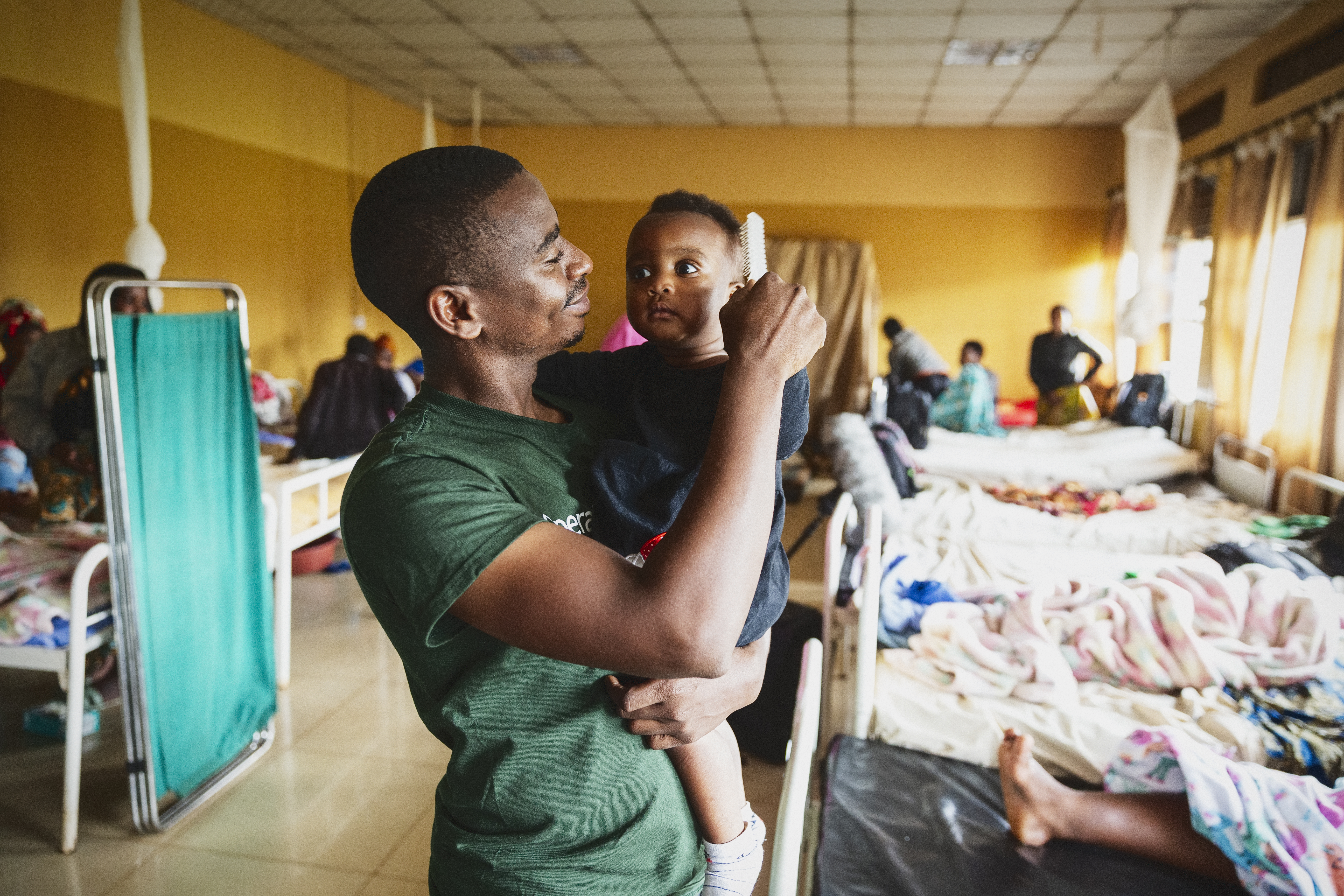 En man håller ett barn i en sjukhussal med flera sängar och personer i bakgrunden.