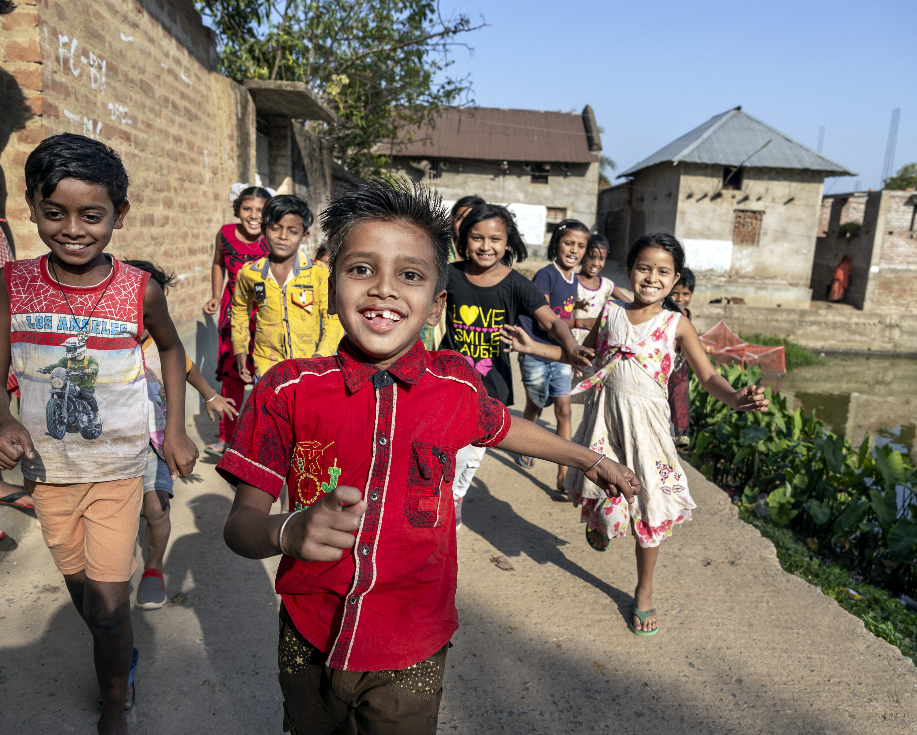 En grupp barn springer mot kameran på en stig i en by, med tegelhus och växtlighet i bakgrunden.