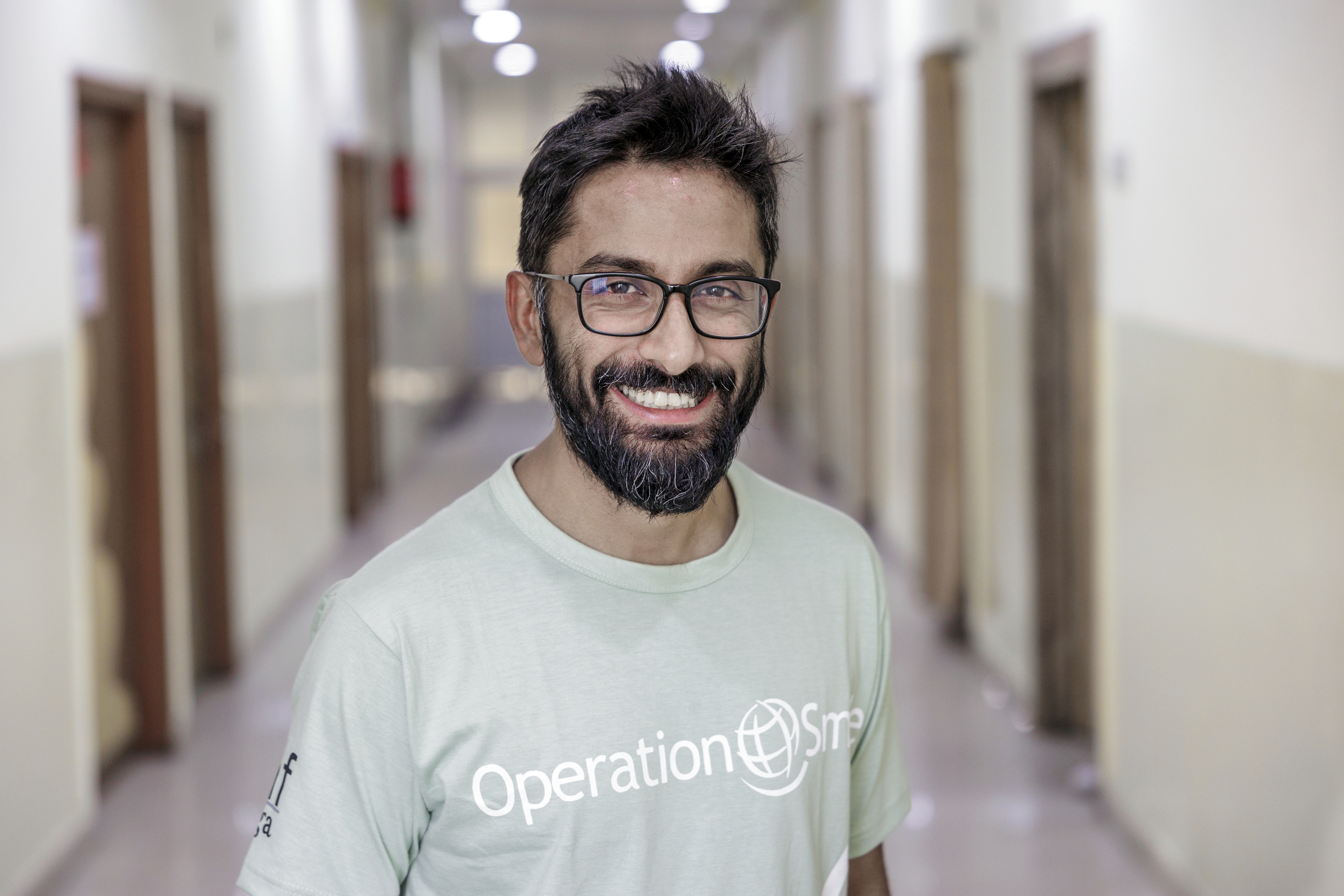 Man med glasögon och skägg, har på sig en Operation Smile t-shirt, står i en korridor med dörrar på båda sidor.