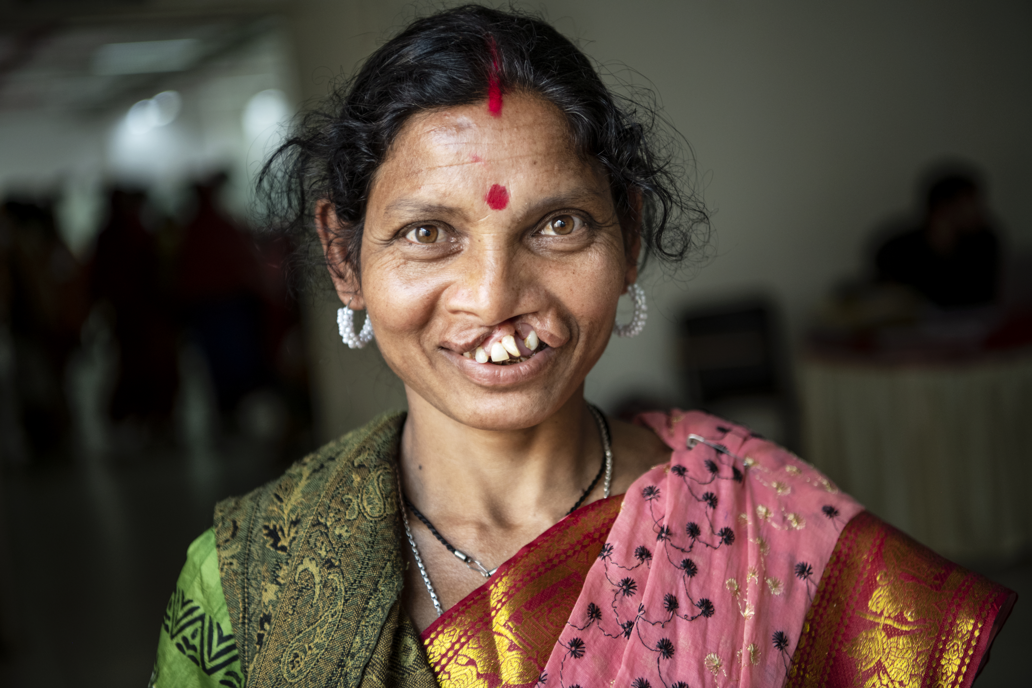 En kvinna med läppspalt ler mot kameran, har grön och rosa sari, traditionella smycken och en röd bindi i pannan.