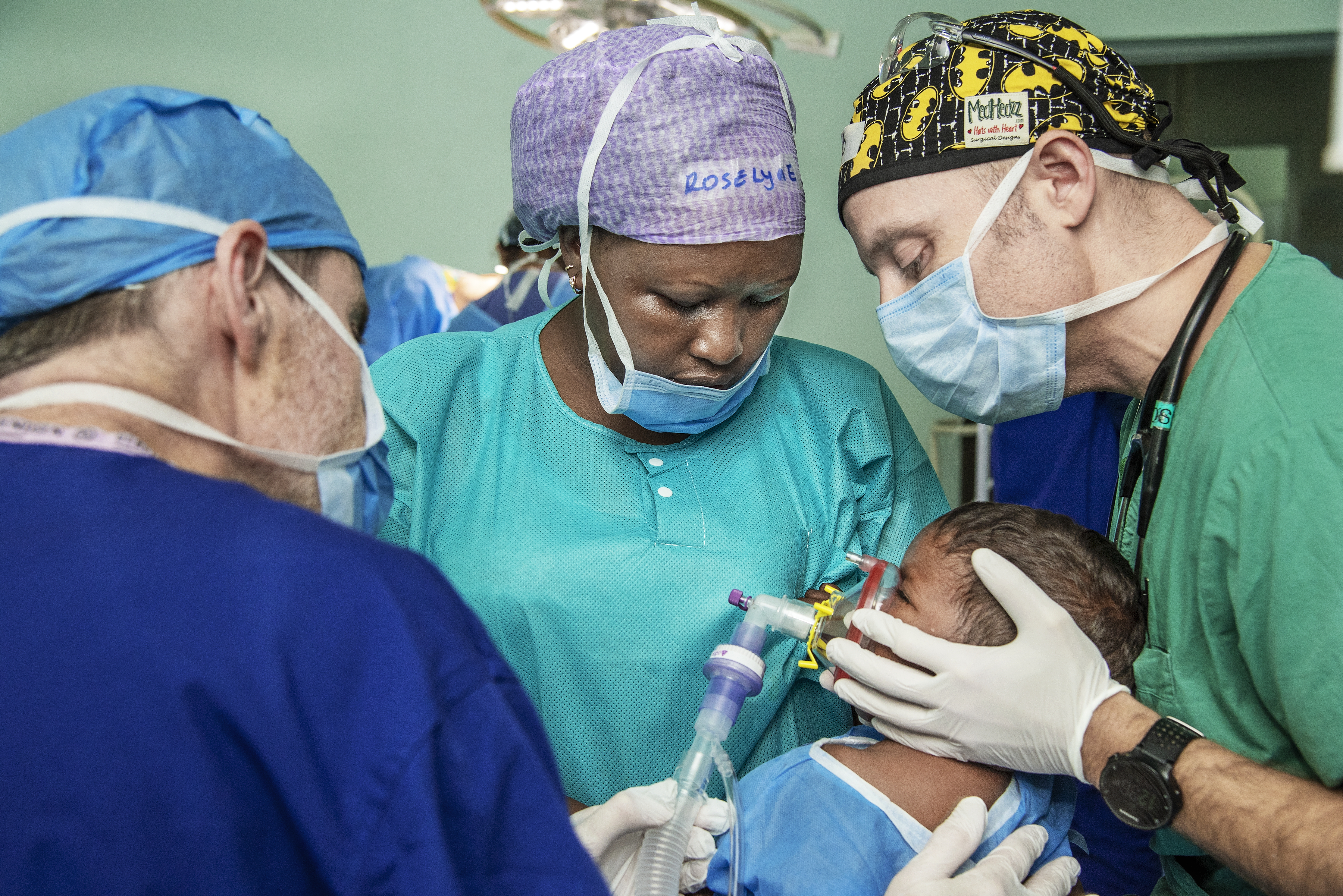 Tre medicinska medarbetare i arbetskläder och munskydd hjälper ett barn som får narkos via en mask i en operationssal.