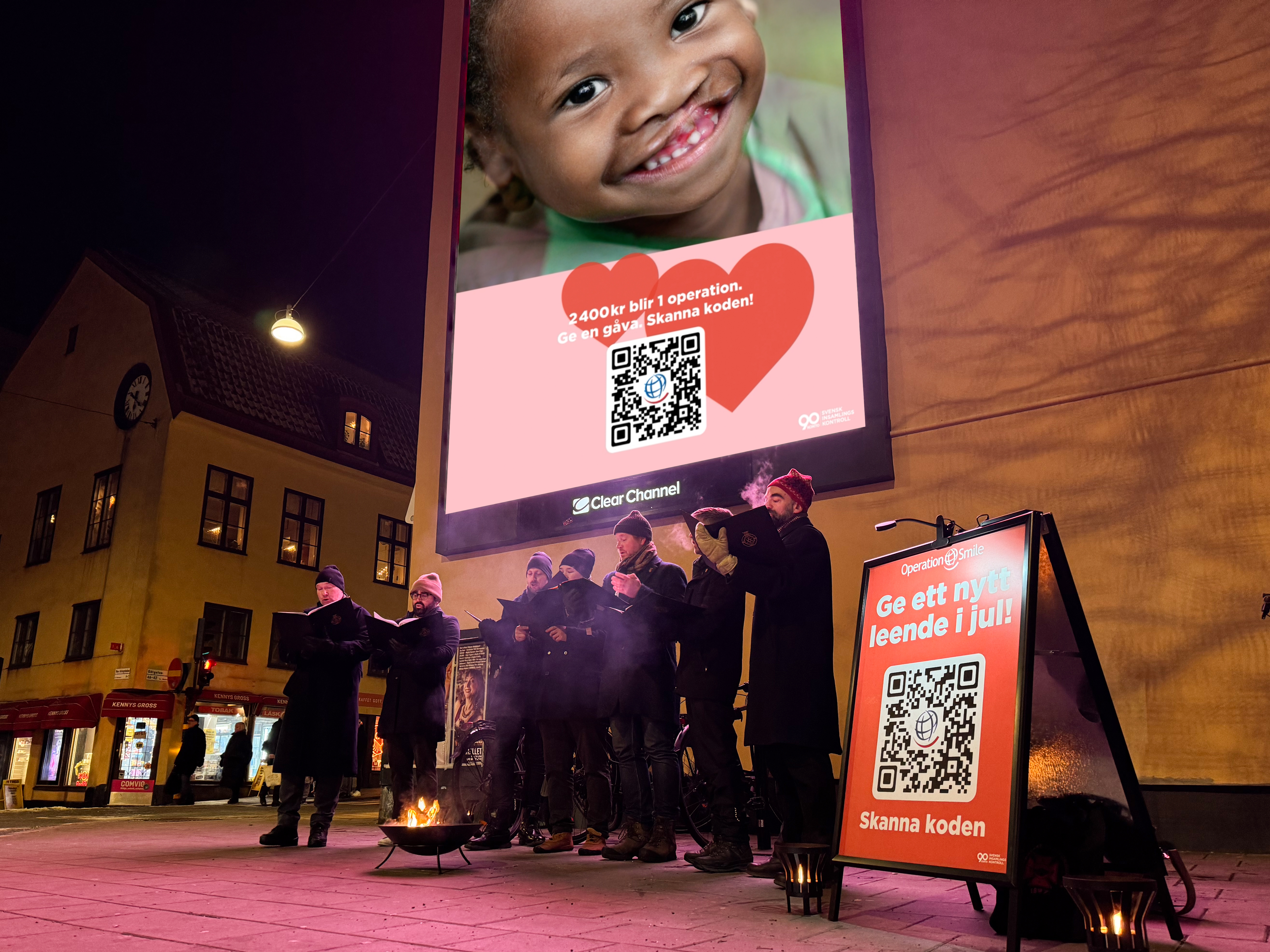 En grupp människor sjunger utomhus på kvällen vid en eldstad under en stor reklamskylt med ett barn och en QR-kod för donationer.