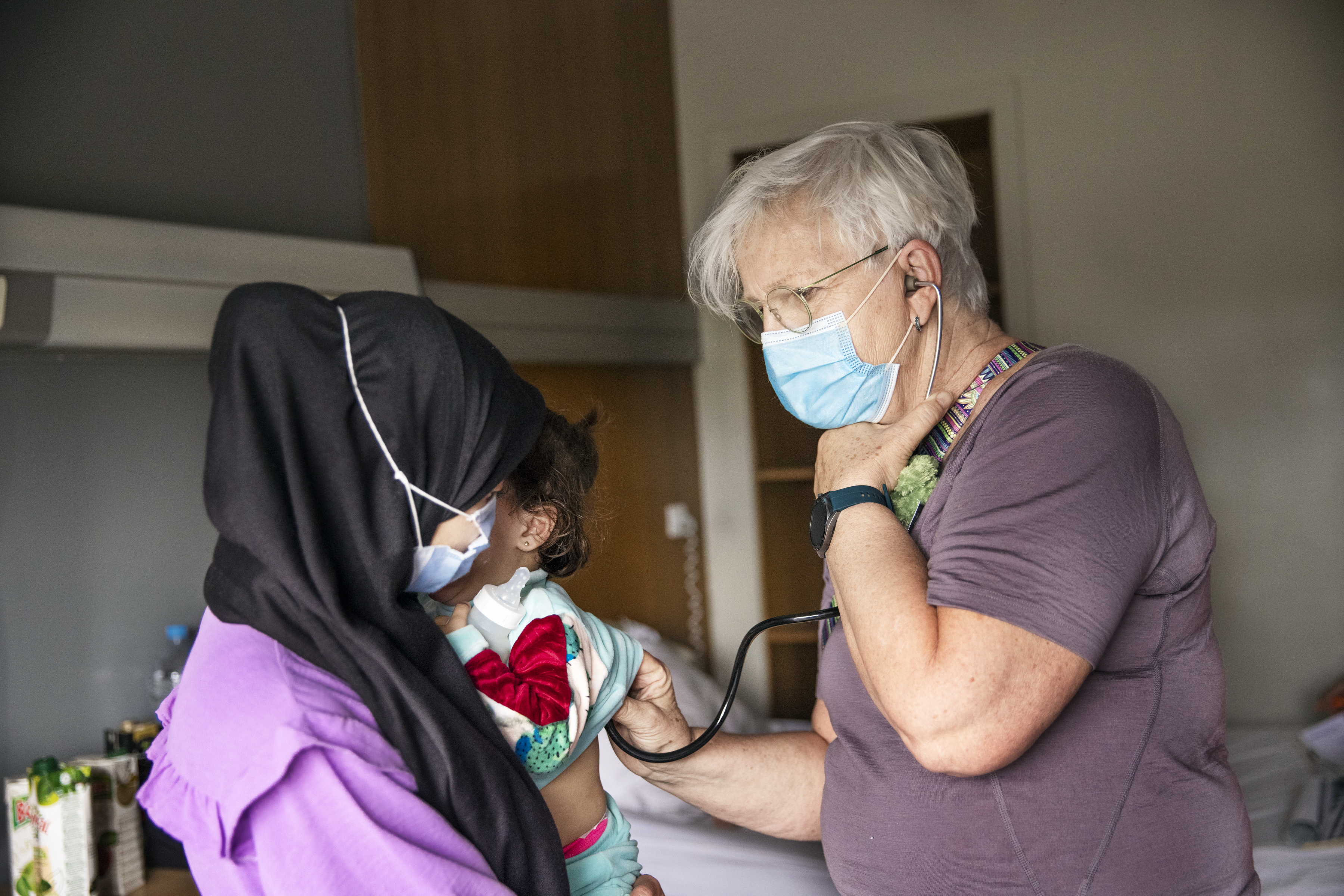En läkare med munskydd använder ett stetoskop för att undersöka ett litet barn som hålls av en kvinna med sjal och munskydd.