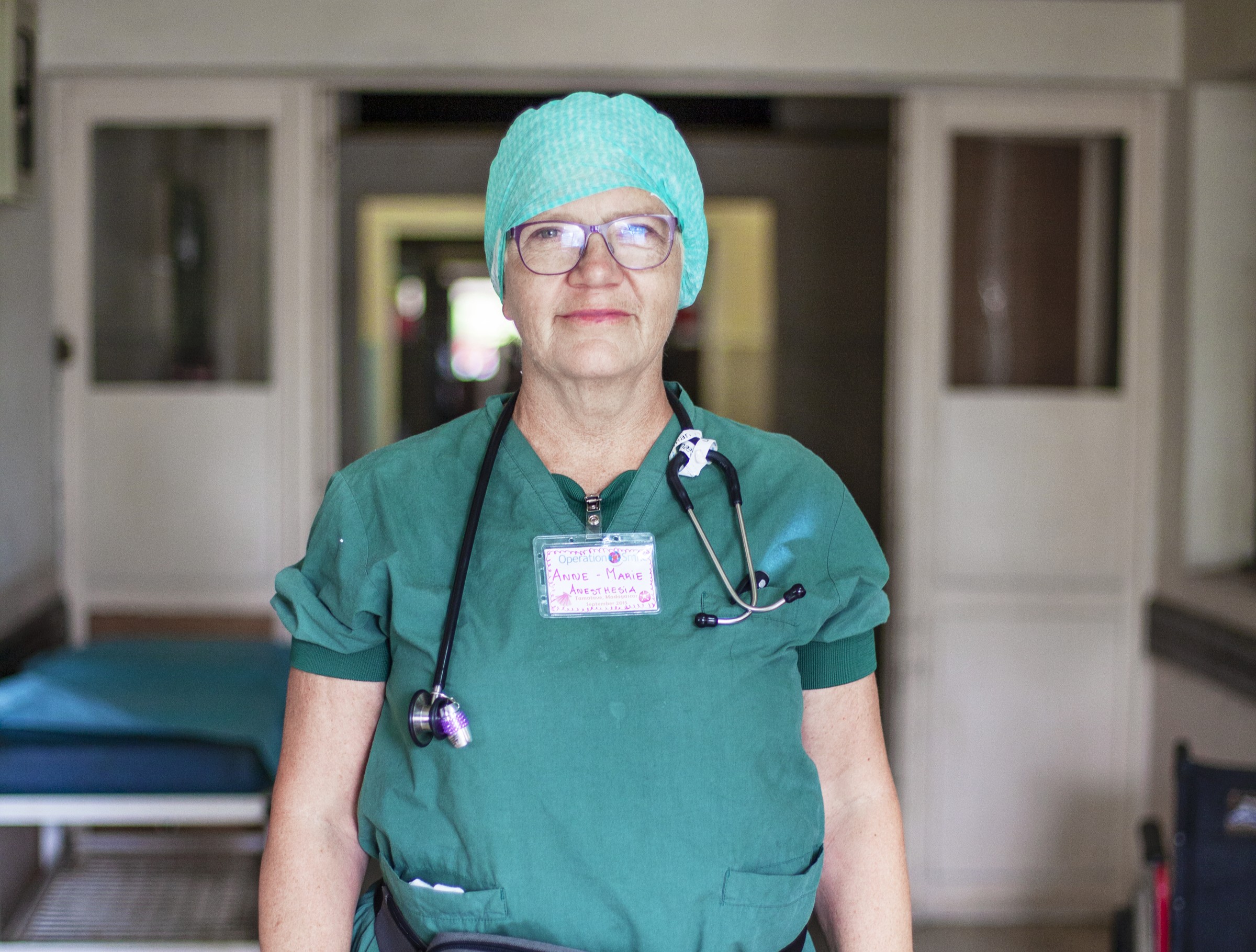 En vårdarbetare i gröna arbetskläder, hårskydd och glasögon står i en sjukhuskorridor med stetoskop och namnbricka.