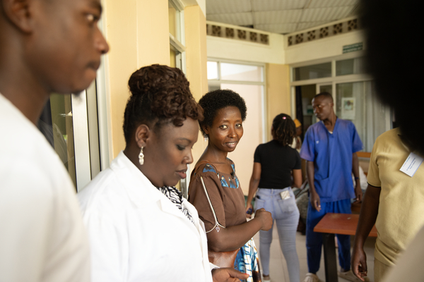 Flera personer, däribland medicinsk personal i vita rockar och arbetskläder, står och talar i en sjukhuskorridor. En kvinna i mitten tittar mot kameran.