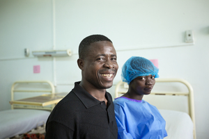 Pappan i svart skjorta tittar mot kameran, medan dottern, nyopererad, är iklädd operationskläder och hårnät. De sitter på en säng i en sjukhussal.