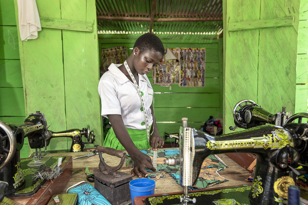En ung kvinna i vit skjorta och grön kjol mäter tyg vid ett bord med symaskiner i ett grönmålat rum.