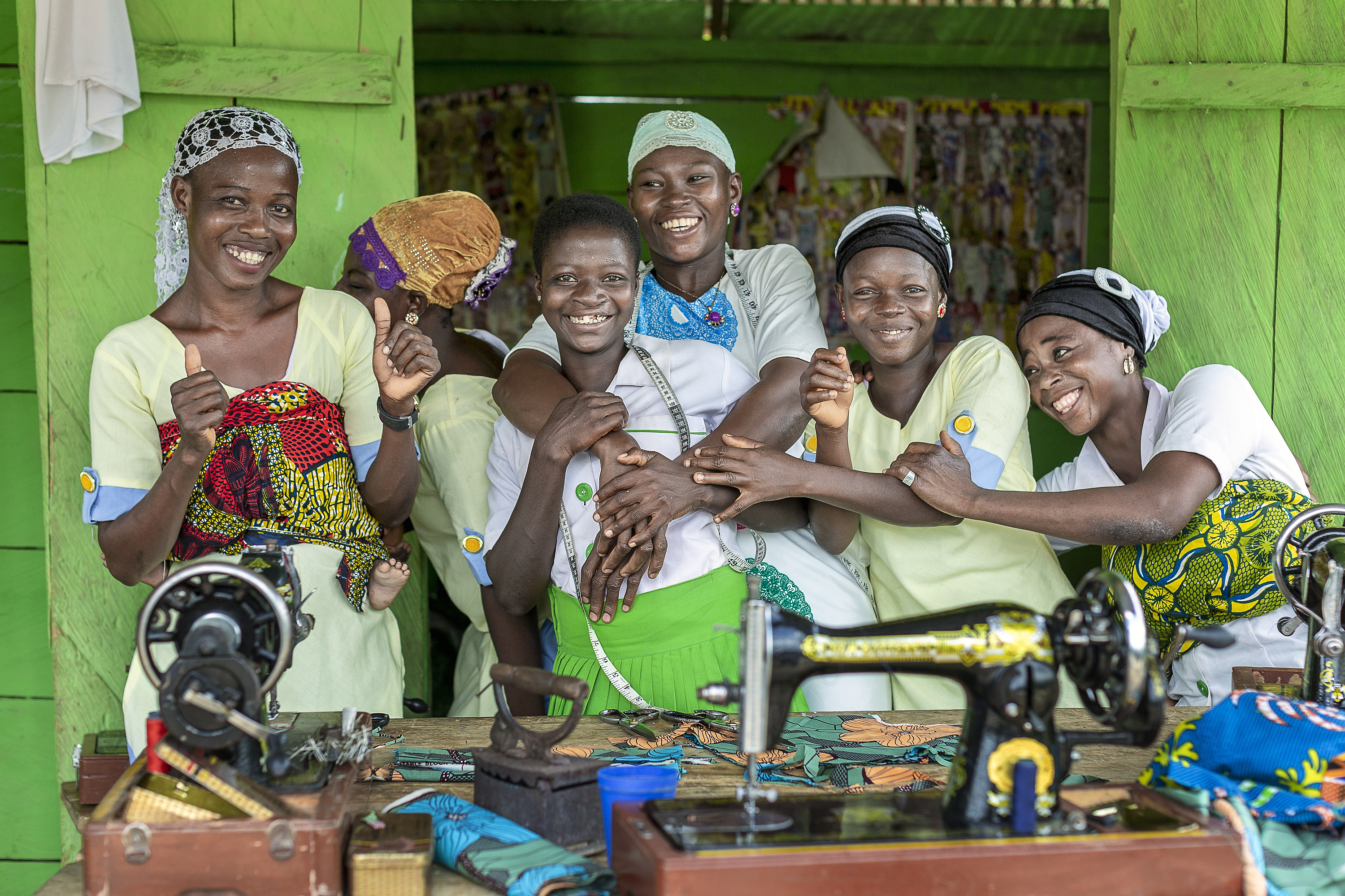 Sex kvinnor ler och poserar tillsammans i en grön syateljé bakom symaskiner och tyg.
