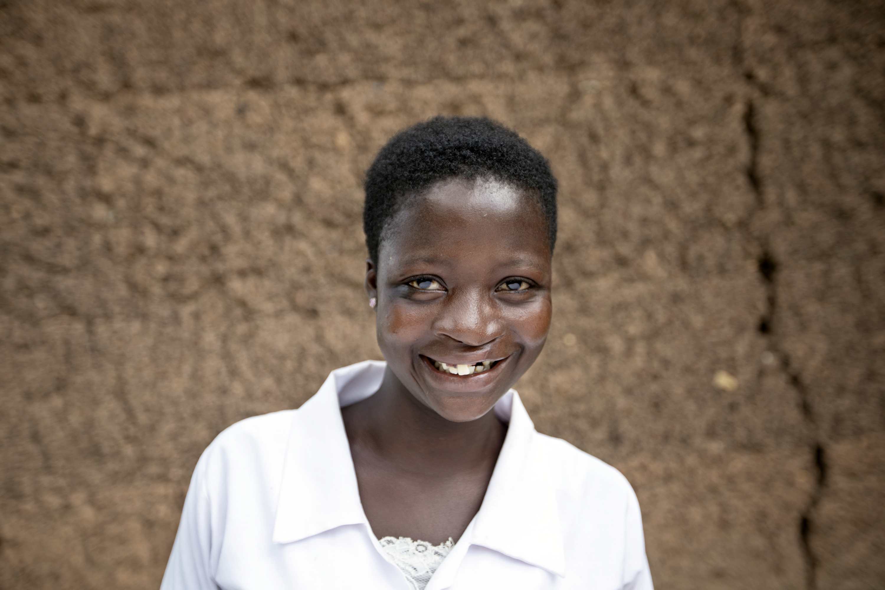 En ung kvinna med kort hår och vit skjorta ler mot kameran och står framför en brun vägg. Hon ser mycket glad ut.