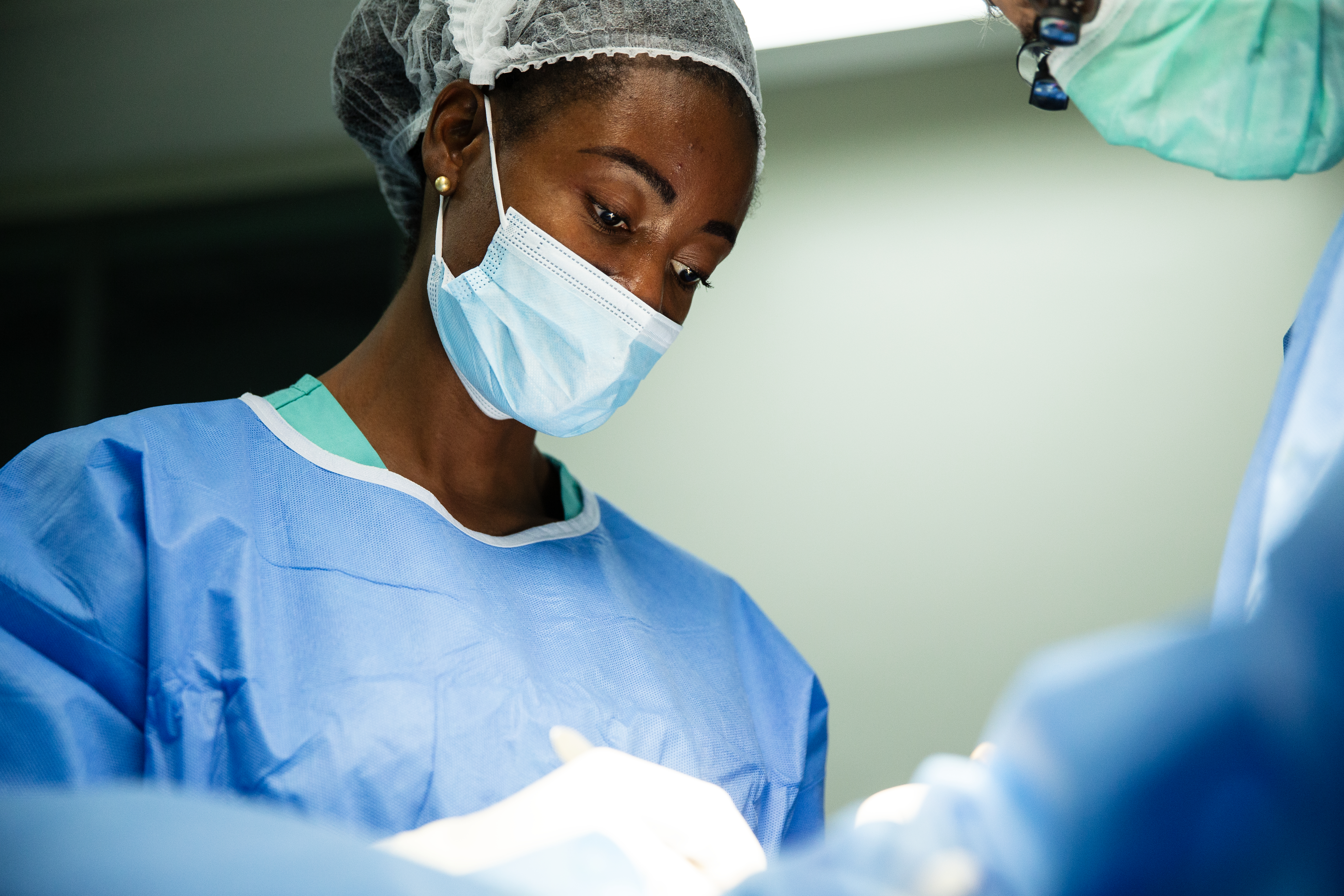 En kirurg med blå rock, munskydd och hårskydd utför ett ingrepp i ett upplyst operationsrum.